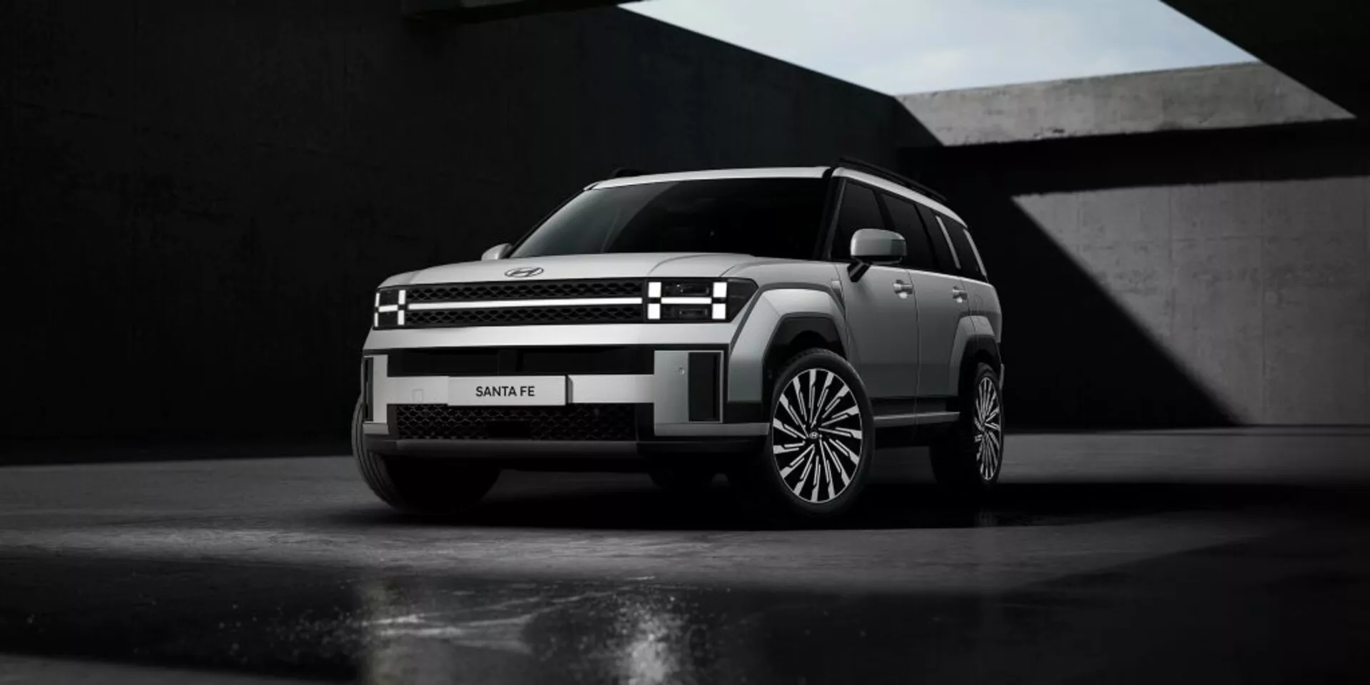 Hyundai Santa Fe 2024 (5. generace) fotografovaný z nízkého úhlu, který zdůrazňuje jeho dominantní postoj a výšku. Obrázek zachycuje levý přední čtvrtinový pohled na vůz, odhalující elegantní přední design, moderní světlomety a mřížku chladiče, stejně jako aerodynamické kontury kapoty. Výrazná vícepaprsková kola a neutrální matná barva šedé nebo stříbrné podtrhují jeho prémiový design. Dramatické osvětlení zvýrazňuje geometrické tvary vozidla, zasazené do minimalistického pozadí, pravděpodobně určeného pro propagační účely.