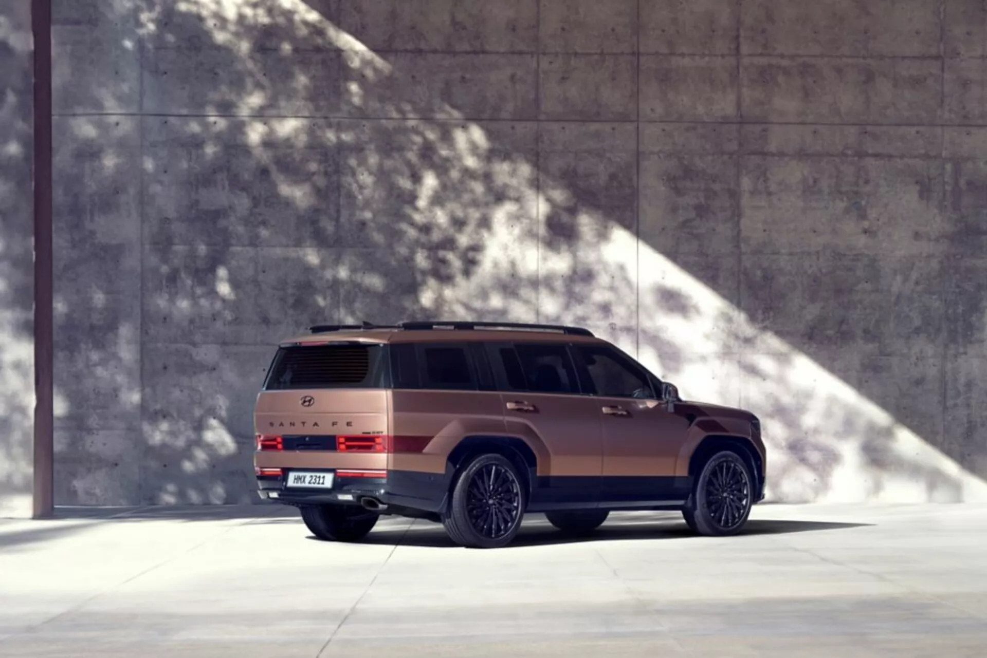 Obrázek Hyundai Santa Fe 2024 (5. generace) v dobře osvětleném prostředí se silnými stíny, zachycený ze tříčtvrtečního pohledu zezadu. Tento moderní SUV je zobrazen z nízkého úhlu, což zdůrazňuje jeho dominantní a robustní vzhled. Vůz je umístěn vlevo, což naznačuje pohyb směrem doprava. Viditelné části zahrnují celý profil pravé strany, zadní část se zřetelnými koncovými světly, zadní nárazník, střešní spoiler a označení. Vozidlo zabírá značnou část rámce a přirozené světlo vrhá ostré stíny napravo, což vytváří prémiový vzhled s matným bronzovým nebo zemním odstínem. Městské nebo architektonické prostředí posiluje moderní design vozidla. Funkce, které lze odvodit, zahrnují litá kola, boční stupínky, střešní okno, LED koncová světla a možné pokročilé technologie, jako jsou parkovací senzory a kamery.