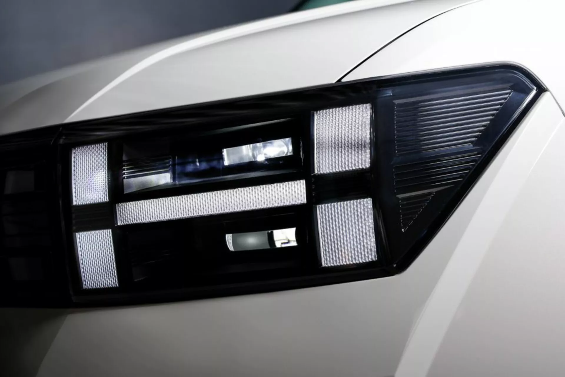 Detailní záběr na moderní světlometový klastr z modelu Hyundai Santa Fe 2024 (5. generace) s pokročilou LED/HID technologií osvětlení, elegantním designem, projektorovou čočkou a lesklým povrchem karoserie.