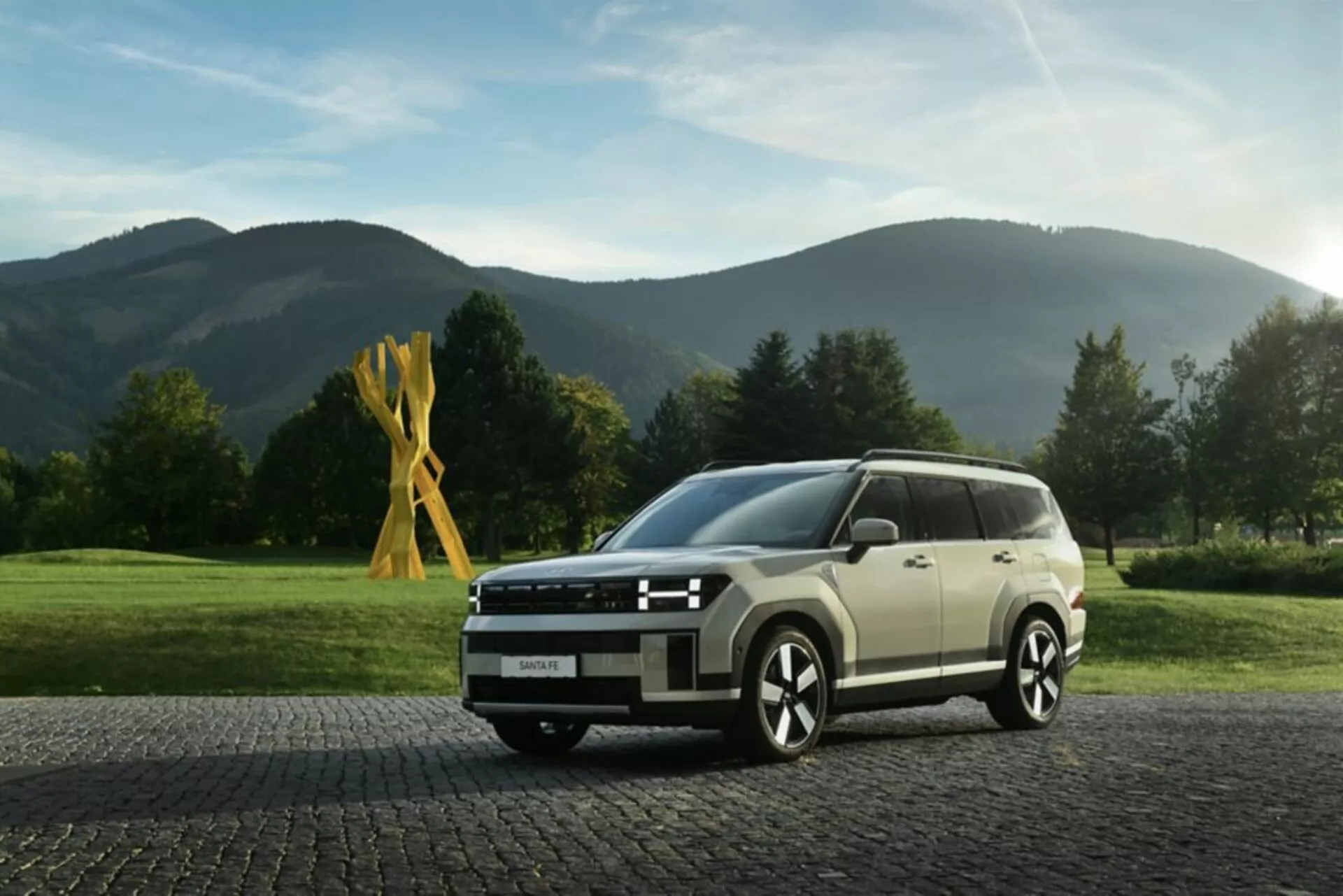 Obrázek zachycuje Hyundai Santa Fe 2024, 5. generace, v nízkoúhlovém záběru z předního kvadrantu, který zdůrazňuje moderní a hranatý design SUV. Viditelné prvky zahrnují LED světlomety s ostrým světelným podpisem, širokou přední mřížku s horizontálním pruhovým designem, částečně viditelný nárazník se senzory nebo kamerami, vícepaprsková litá kola pro sportovní vzhled a boční zrcátka s integrovanými směrovými světly. Linie karoserie podél strany zdůrazňují celkový designový jazyk vozidla, zatímco velká okna mohou mít UV filtraci. Auto je nastaveno na dlažebních kostkách s klidným, venkovským pozadím, což zvyšuje jeho všestrannou přitažlivost pro městské i venkovské prostředí. Osvětlení a hloubka ostrosti zaměřují pozornost na vozidlo, což ho činí vhodným pro kategorie jako moderní SUV, automobilový design a luxusní vozidla.