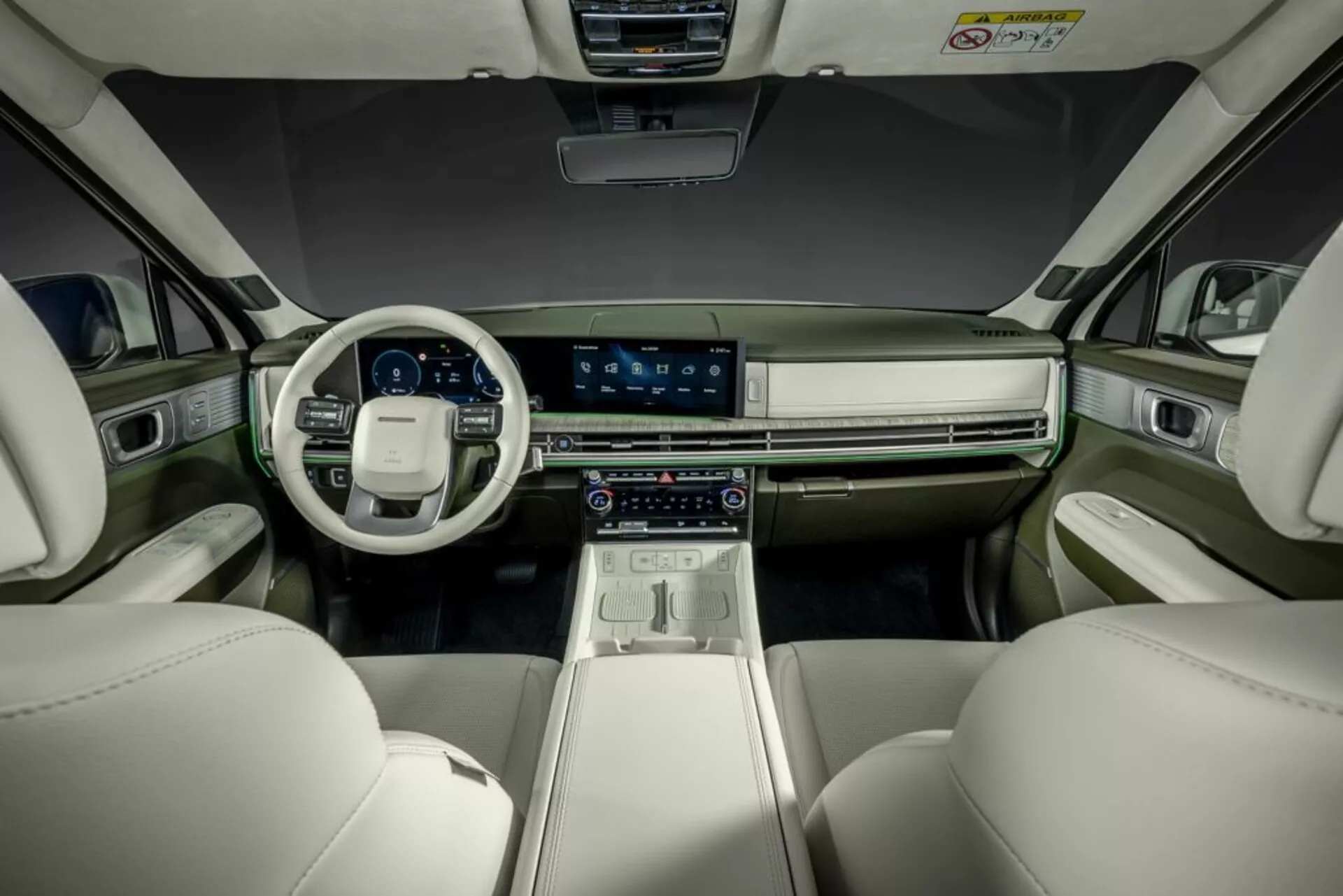 Interiérová fotografie Hyundai Santa Fe z roku 2024 (5. generace), zachycující pohled ze zadního sedadla směrem k palubní desce. Obrázek zdůrazňuje přední sedadla a kokpit, zahrnující výrazný centrální dotykový displej, digitální přístrojovou desku, pokročilé ovládání klimatizace a volant s integrovanými ovládacími prvky. Moderní, luxusní kabina má dvoubarevné barevné schéma s světlými koženými sedadly a tmavší palubní deskou a dveřmi, doplněné kovem. Atmosféru vylepšuje ambientní osvětlení.
