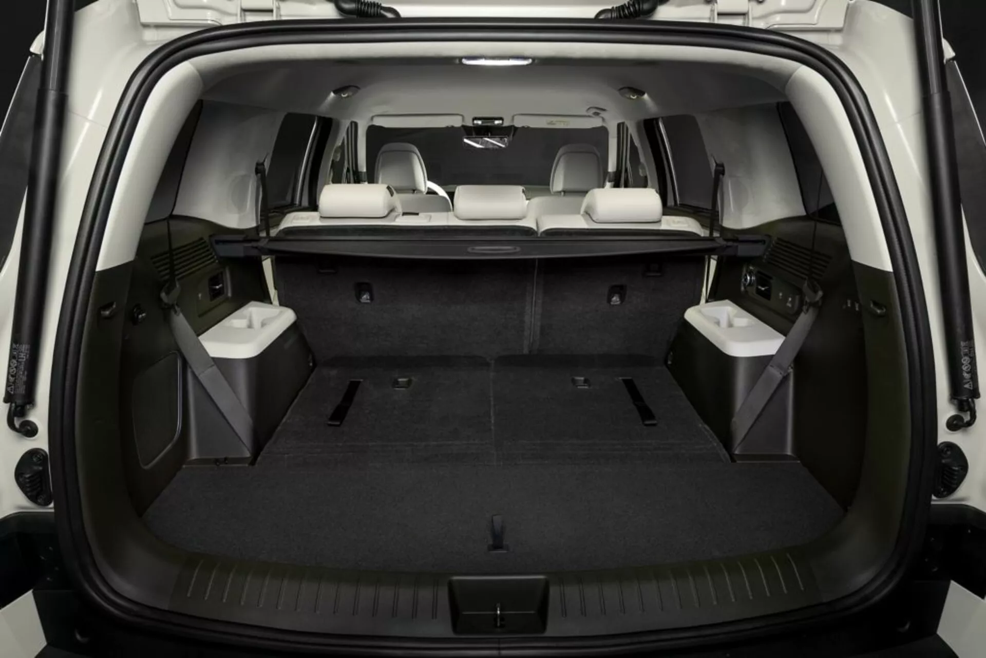 Obrázek zobrazuje interiér nákladového prostoru Hyundai Santa Fe 2024 z pohledu zezadu, s otevřenými pátými dveřmi, které odhalují nepoškozenou, plochou nákladovou plochu pokrytou kobercem. Třetí řada sedadel je sklopena pro rozšíření nákladového prostoru, zatímco prostřední řada zůstává vzpřímená pro cestující. Dobře osvětlený interiér zvýrazňuje prvky jako úchyty pro upevnění, boční úložné přihrádky a integrované panely, pravděpodobně obsahující reproduktory nebo větrací mřížky.