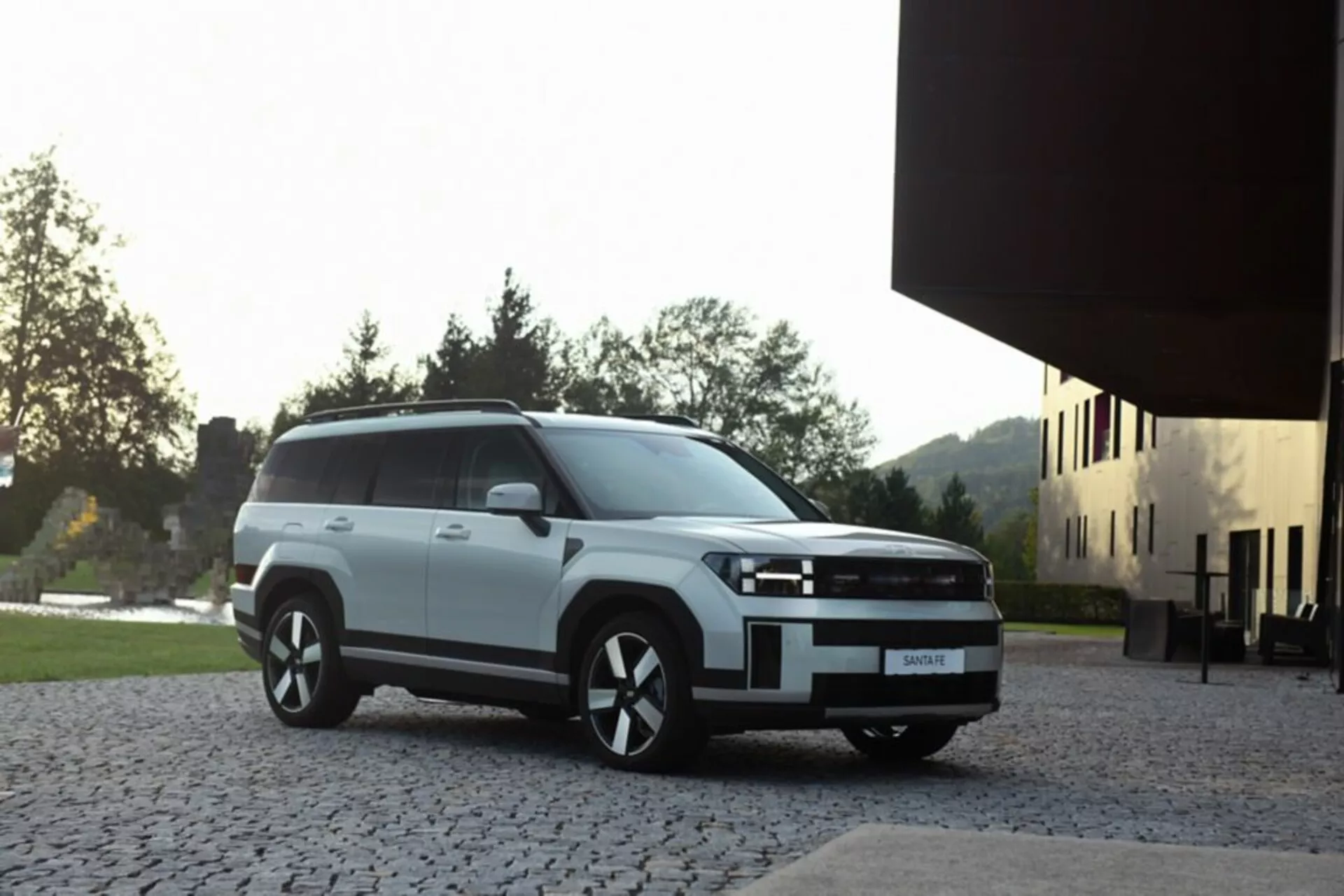 Hyundai Santa Fe 2024 5. generace v bílé barvě, zaparkovaný na kamenném povrchu s moderní budovou a zelení v pozadí, zobrazený z předního tříčtvrtečního úhlu, se čtvercovou přední maskou, LED nebo HID světlomety a litými koly, osvětlený měkkým přirozeným světlem zvýrazňujícím moderní estetiku SUV.