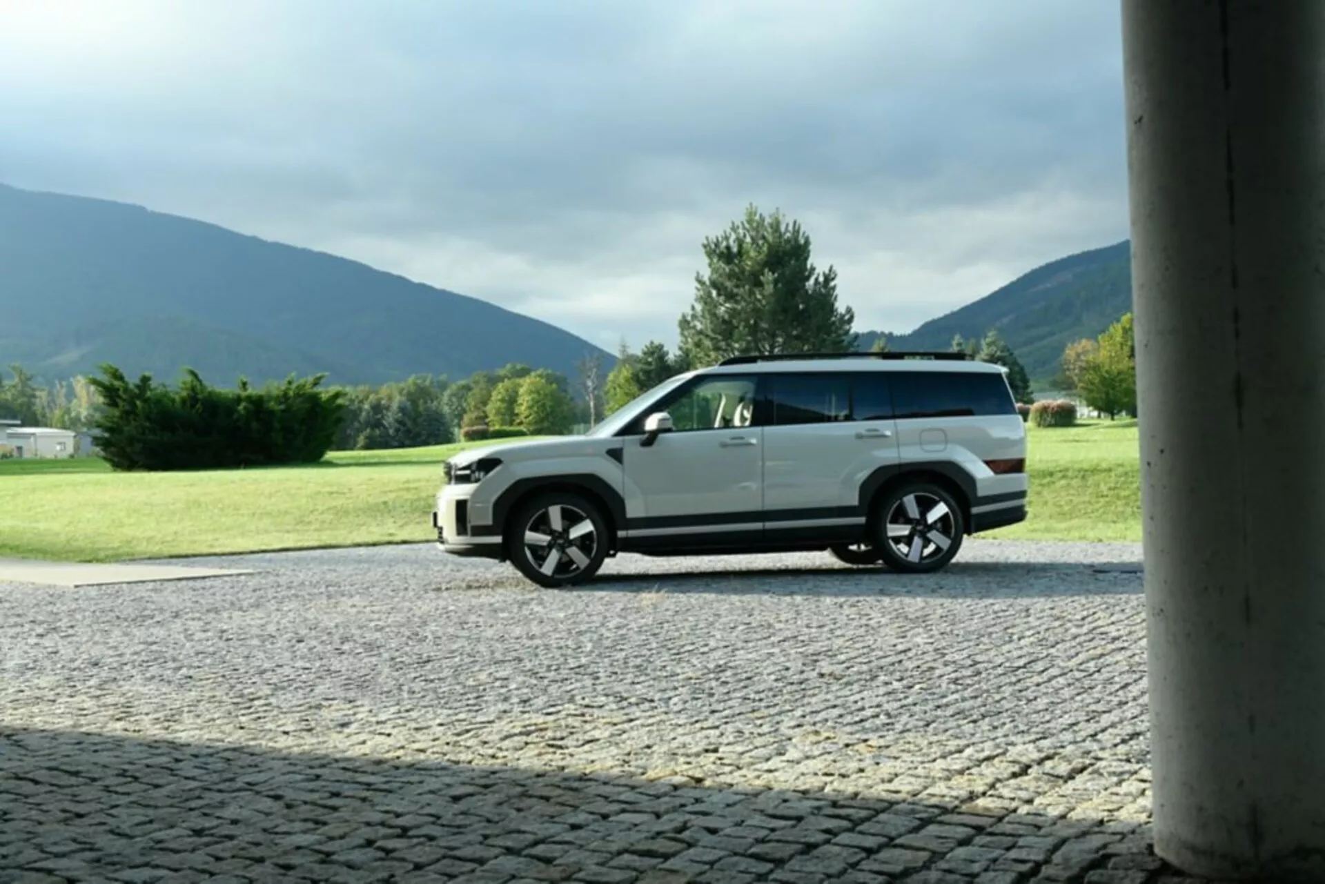 Boční profil vozu Hyundai Santa Fe 2024 (5. generace), zaparkovaného na dlážděném povrchu s přírodním pozadím. Obrázek zachycuje moderní design vozu, zvýrazňuje velká vícepaprsková kola, dvoubarevné lakování a detaily karoserie. Fotografie, orámovaná sloupem stavby, je pořízena z nižšího úhlu a zdůrazňuje výšku a postoj auta. Venkovní nastavení během dne zvýrazňuje exteriér vozu bez silných stínů.