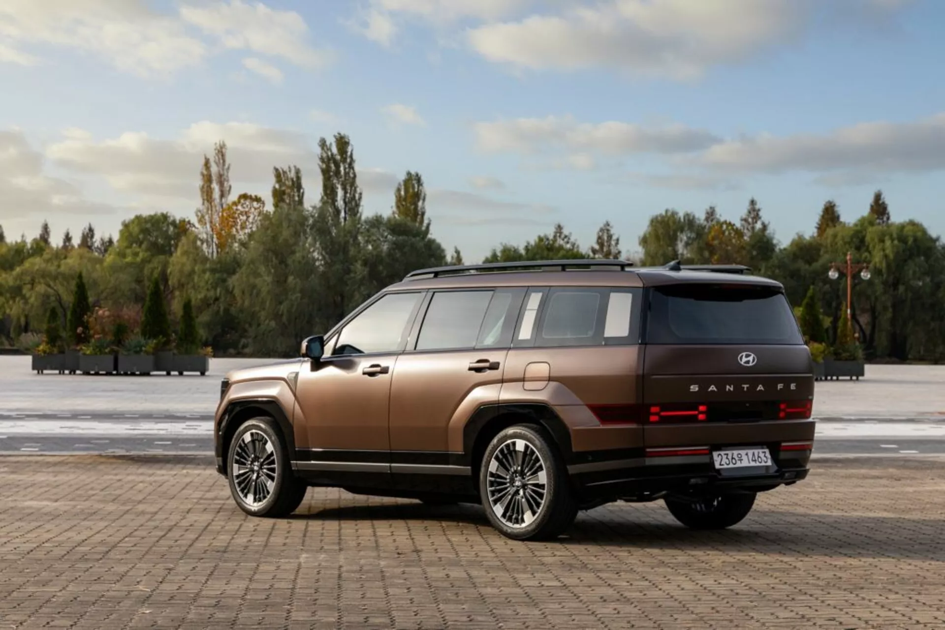 Obrázek zobrazuje moderní SUV Hyundai Santa Fe 2024, 5. generace, na otevřeném dlážděném prostoru obklopeném stromy a modrou oblohou s rozptýlenými mraky. Fotografie pořízena z úhlu tři čtvrtin zezadu ukazuje vysokou linii pásu, ploché boční panely a hranaté blatníky s velkými, vícepaprskovými litými koly. SUV má dvoubarevný lak v měděné hnědé s černým spodním lemem. Zadní část obsahuje vodorovně orientovaná LED zadní světla, stříbrný ozdobný proužek, označení modelu 'SANTAFE' a odrazky na nárazníku. Anténa ve tvaru žraločí ploutve a panoramatické střešní okno naznačují pokročilé technické prvky. Přirozené denní světlo zvýrazňující kontury auta doplňuje scénu.
