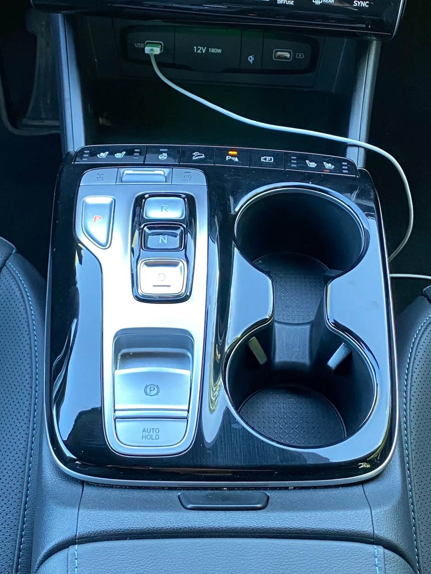 Obrázek zobrazuje centrální konzoli Hyundai Tucson 2020 s elektronickým převodovým voličem ve stavu 'N' obklopeným lesklým černým dekorem. Na levé straně jsou ovládací prvky, včetně tlačítka napájení, voličů jízdních režimů a funkce 'AUTO HOLD'. Vpravo jsou dvojité držáky nápojů, z nichž jeden obsahuje bílý nabíjecí kabel. V horní části je vidět část infotainmentového displeje s indikátory USB portů, což ukazuje na moderní technologie a luxus vozu.