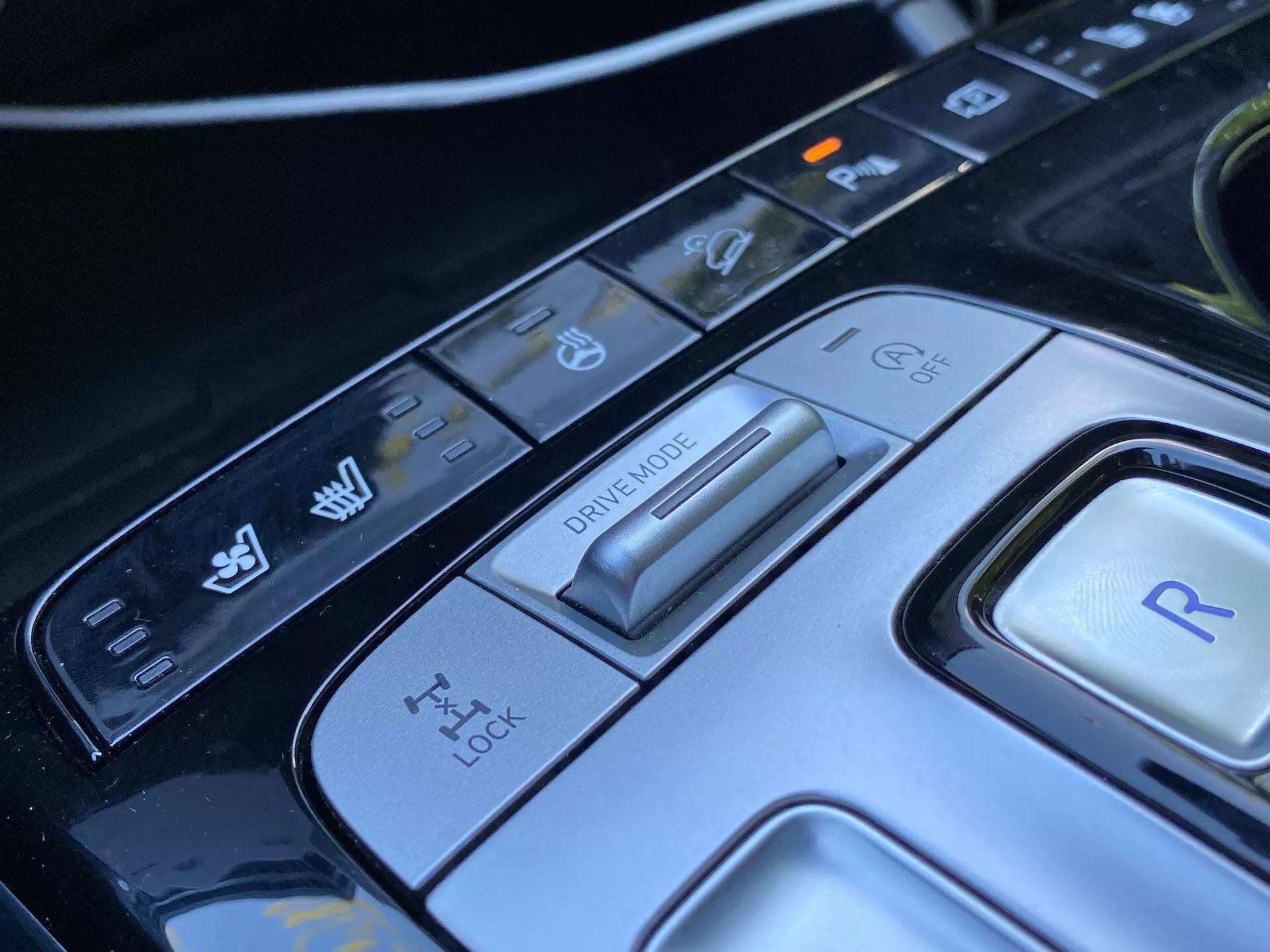 Detailní pohled na středovou konzoli vozu Hyundai Tucson 2020, zaměřený na volič jízdního režimu a různé ovládací tlačítka. Obraz ukazuje pokročilý přepínač jízdního režimu pro různé jízdní podmínky, panel pro kontrolu trakce, tlačítko pro zpátečku a další ovládací prvky pro stabilitu vozidla a asistent parkování. Elegantní design a high-tech materiály zdůrazňují sofistikovaný automobilový interiér tohoto luxusního moderního auta.