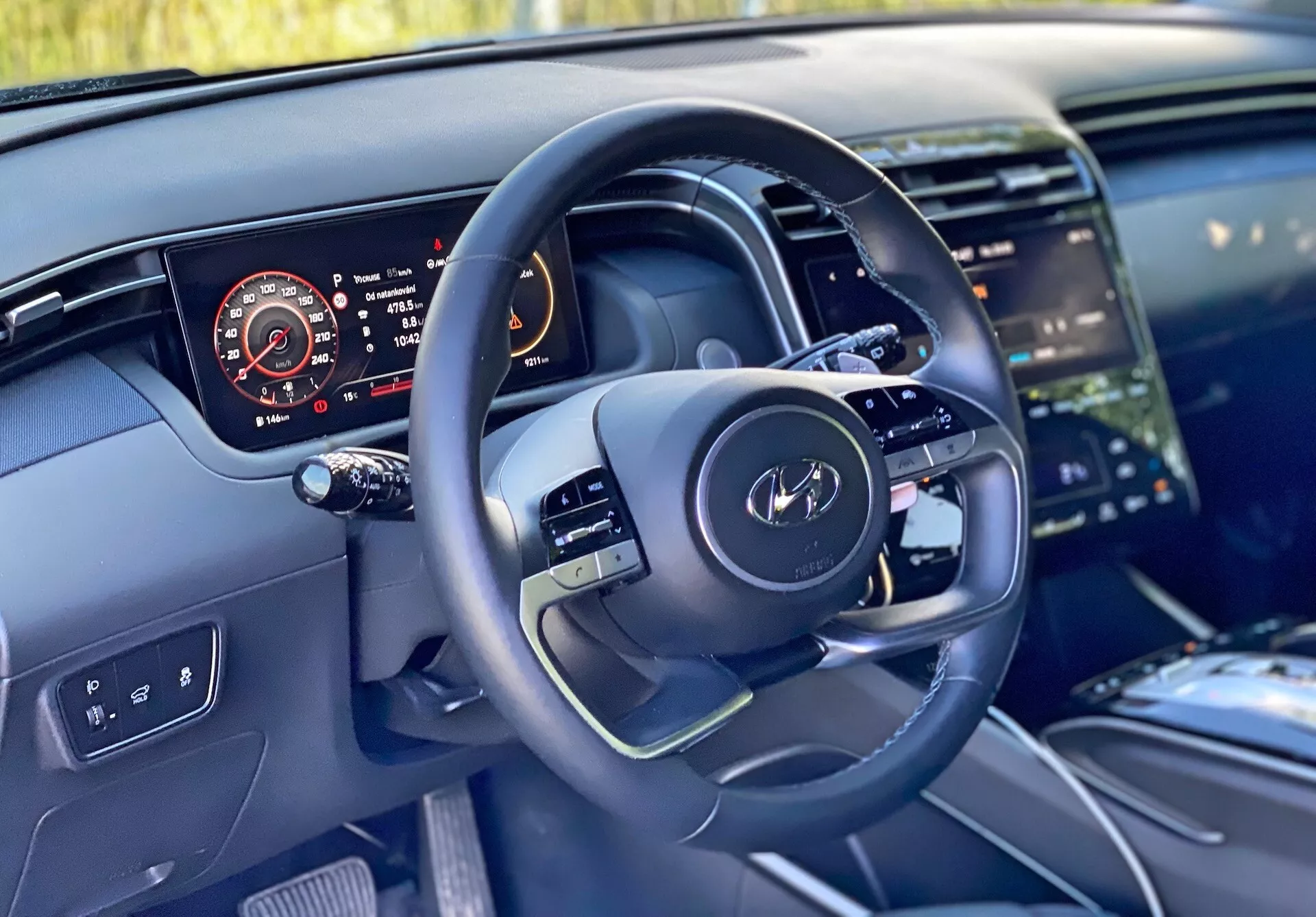 Interiér vozu Hyundai Tucson 2020 z pohledu řidiče ukazující moderní volant s multifunkčními ovladači a digitální přístrojový panel zobrazující rychloměr, otáčkoměr, hladinu paliva a teplotu motoru. Obrázek také zdůrazňuje část středového panelu s velkým dotykovým infotainment systémem, čistý design palubní desky s ambientním osvětlením a vysoce kvalitními materiály naznačujícími luxusní povrchy.