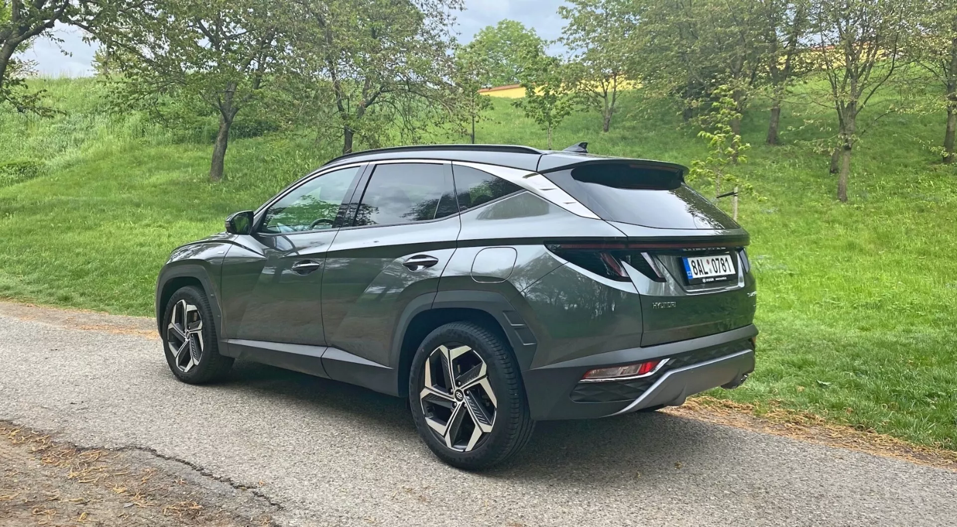 Pohled na tři čtvrtiny zezadu na 2020 Hyundai Tucson 1.6 T-GDi Pure SUV zaparkovaný na úzké silnici v venkovském prostředí, který ukazuje velká vícepaprsková litá kola, tmavý metalický lak s klesající linií střechy, zadní spoiler, zatmavená okna vzadu, moderní zadní světla a dvojité výfukové koncovky integrované do zadního nárazníku.