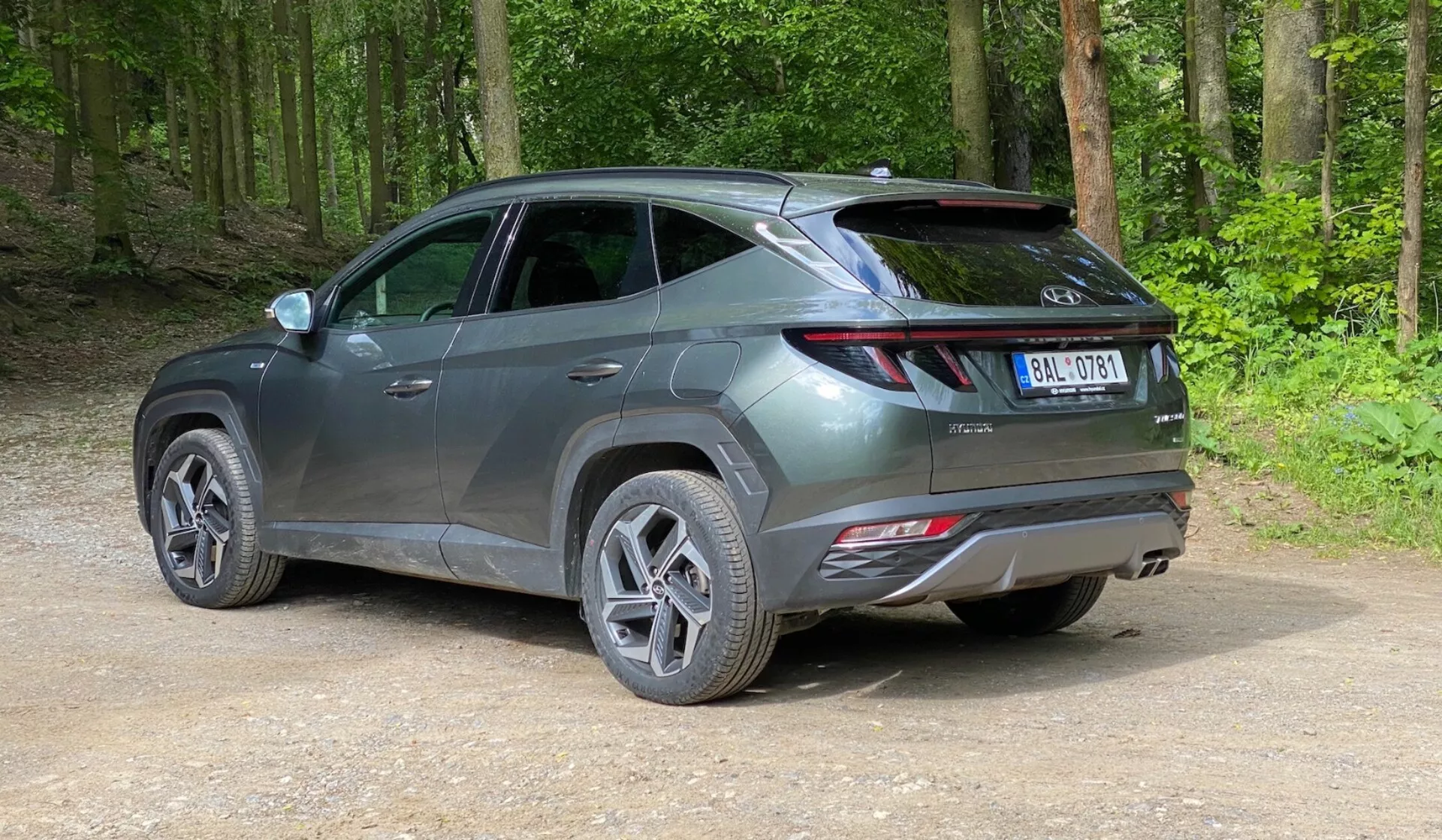 Moderní SUV zaparkované na neudržované cestě v lesní oblasti, zachyceno z zadního tříčtvrtečního pohledu z mírné elevace. Vozidlo má elegantní, současný design s ostrými liniemi, protáhlými světly obepínajícími ze zadní strany na boky, viditelným spoilerem a reflektory na zadním nárazníku, což naznačuje pokročilé bezpečnostní funkce. Vozidlo v metalické šedé barvě má vícepaprsková litá kola a silniční pneumatiky, lesklý povrch naznačuje, že je nové nebo dobře udržované. Okolo auta je zelený, bujný les s ušlapaným bahnem a uvolněným štěrkem na zemi.