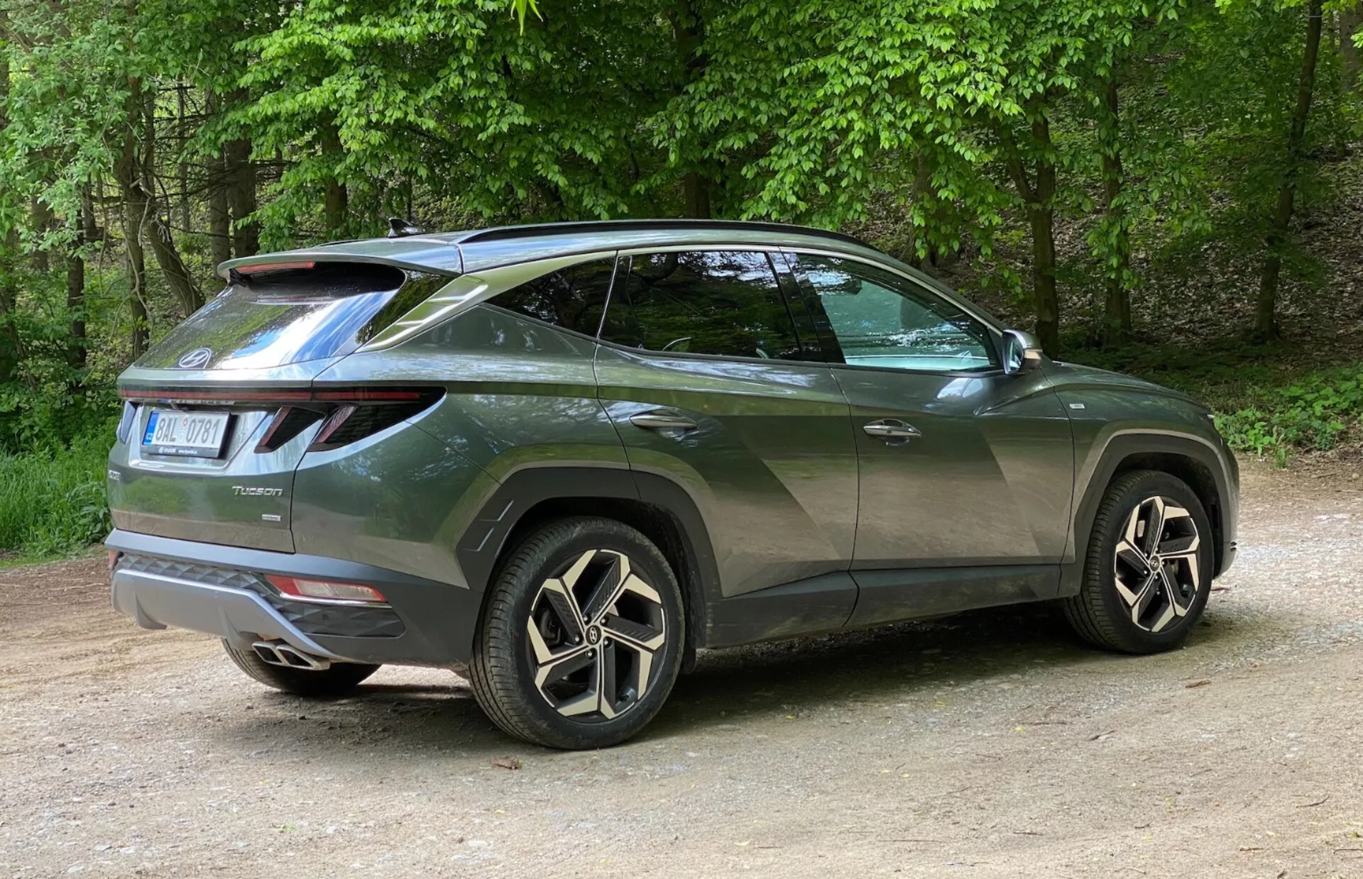Zadní pohled na moderní SUV Hyundai Tucson 2020 se zaměřením na boční profil a zadní design. Vozidlo je umístěno vlevo, fotografie je pořízena zprava, zdůrazňujíc zadní pravý roh. Charakteristická LED zadní světla se obklopují od zadní části k bokům a střešní spoiler dodává sportovní vzhled. Stoupající linie oken, šikmá střecha, tónovaná okna a kliky dveří v barvě karoserie zvýrazňují sportovní vzhled. Velká hliníková kola s více paprsky a nízkoprofilové pneumatiky zvyšují luxusní vzhled. Drobnosti jako zadní stěrač a kryty blatníků znamenají praktičnost a robustnost. Auto je umístěno na štěrku v přírodním prostředí se stromy v pozadí.