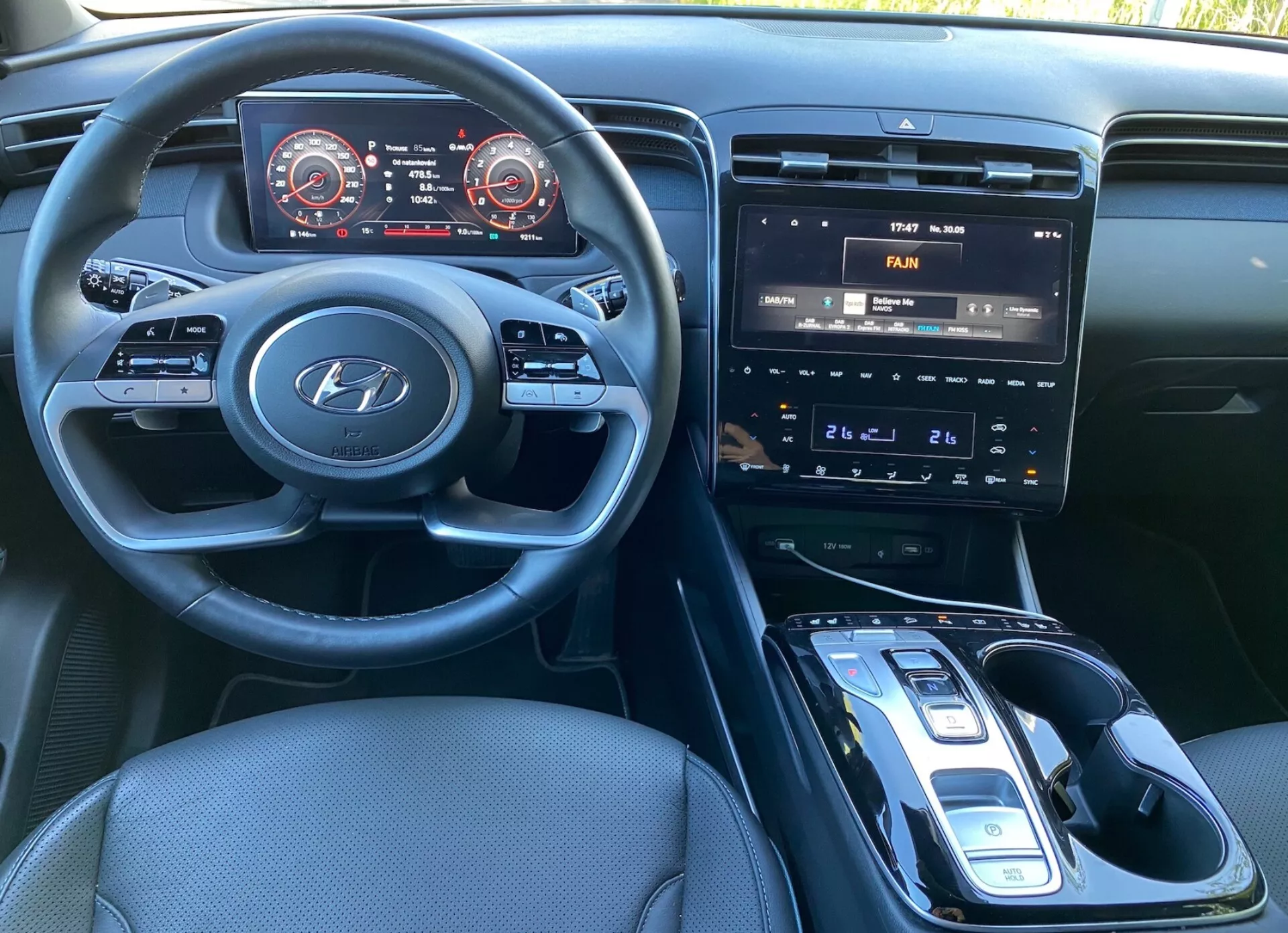 Pohled na interiér Hyundai Tucson 2020 ze sedadla řidiče, ukazující volant s logem Hyundai a ovládacími prvky pro audio, telefon a tempomat. Přístrojová deska obsahuje digitální panel s tachometrem a rychloměrem s červeným zvýrazněním. Středová konzola má infotainment systém s dotykovou obrazovkou zobrazující ovládání klimatizace, fyzická tlačítka a knoflíky na ovládání klimatizace a automatický řadicí páku s elektronickým selector arm. Je vidět potenciální bezdrátová nabíječka, tmavé čalounění z textilie a dobře osvětlený interiér.