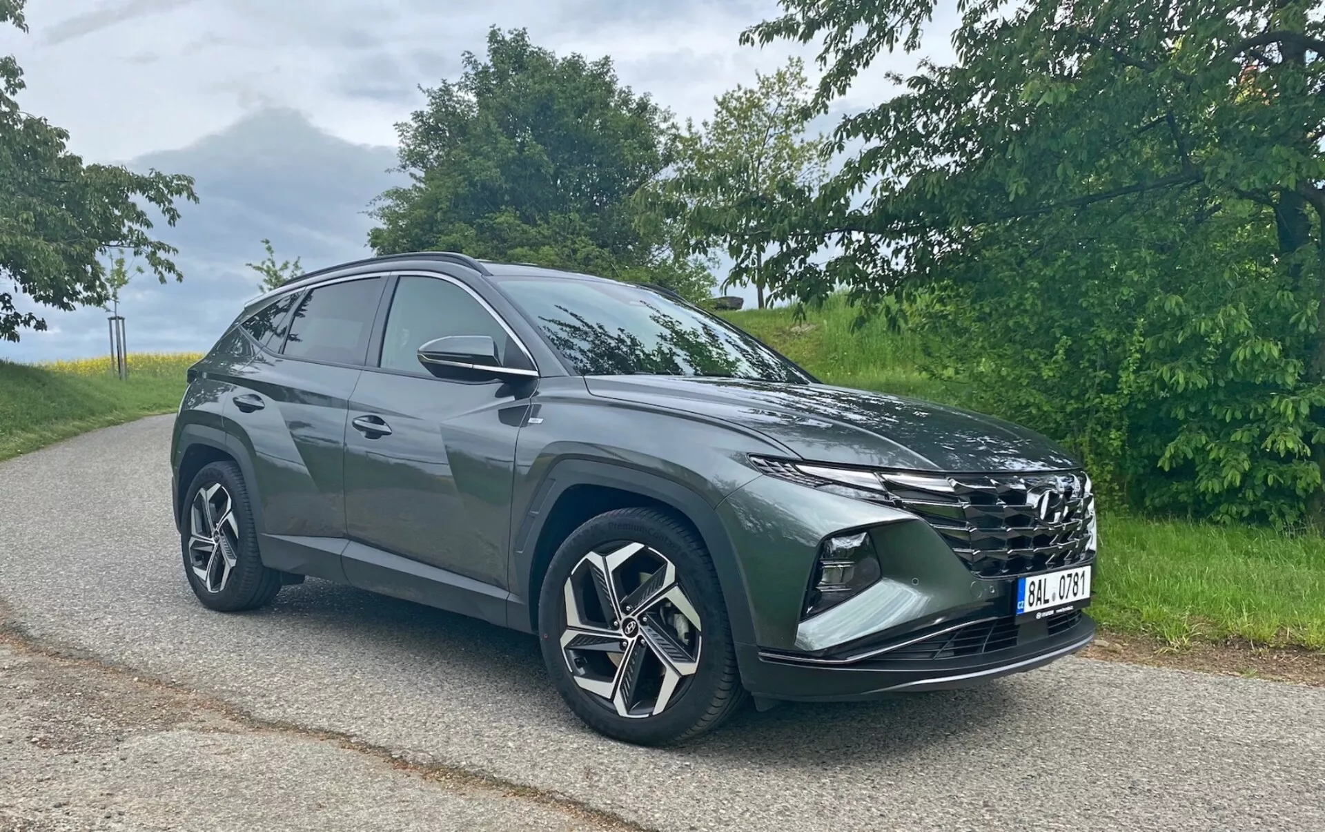 Obrázek zobrazuje moderní SUV Hyundai Tucson 1.6 T-GDi Pure zaparkované na asfaltové silnici s přírodním pozadím. Vozidlo je fotografováno z předního tříčtvrtečního úhlu, což zviditelňuje jeho výraznou masku chladiče, elegantní světlomety, přední nárazník s mlhovými světly nebo vstupy vzduchu, velká litá kola s vícepaprskovým designem a zrcátka. Silueta SUV se směrem dozadu sklání, což naznačuje sportovní, aerodynamický design. Snímek byl pořízen za měkkých denních světelných podmínek, což zvýrazňuje barvu a detaily vozidla bez ostrých stínů.