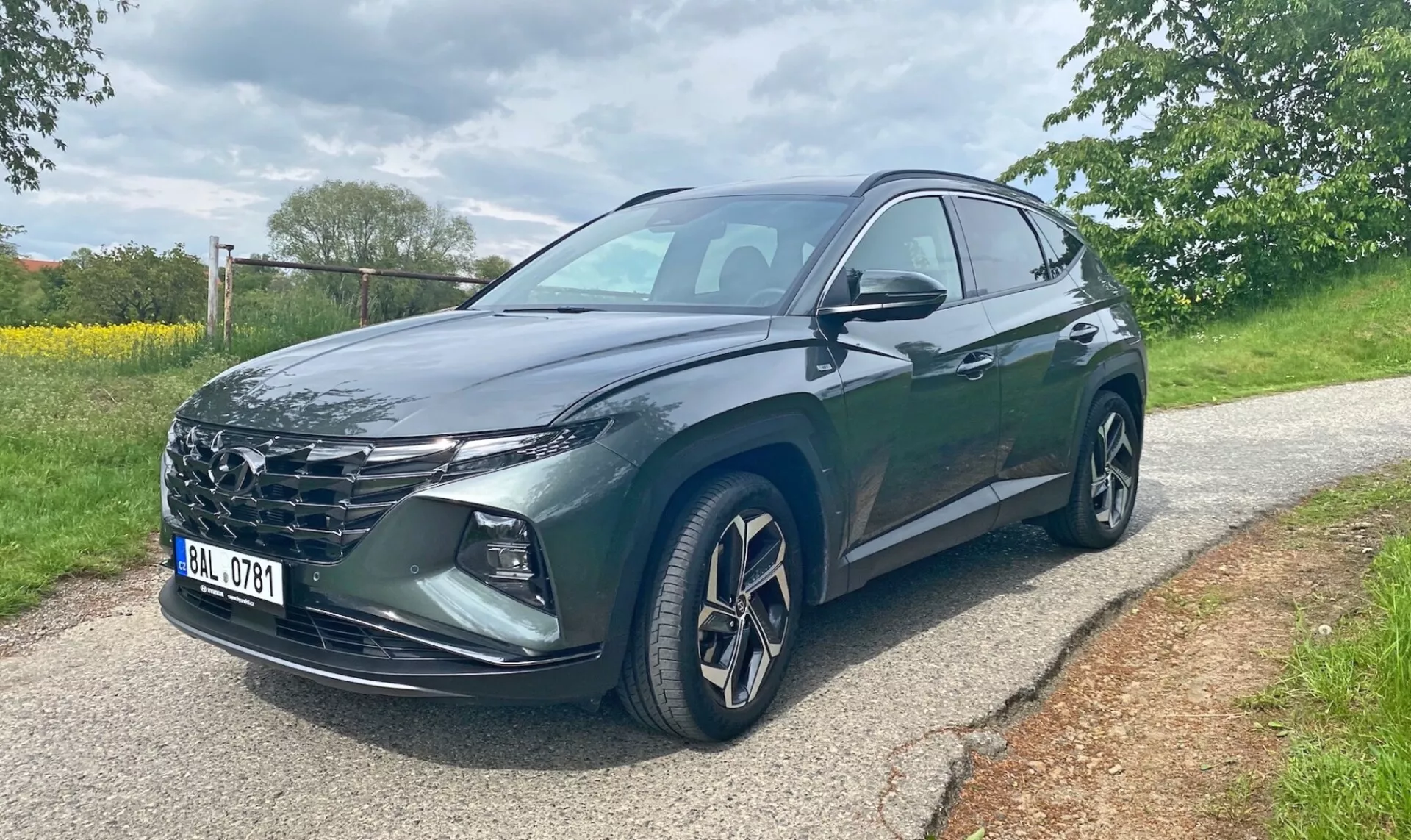 Hyundai Tucson 2020 1.6 T-GDI Pure SUV, zobrazený z předního třičtvrtinového úhlu na dlážděné silnici s stromy a trávou v pozadí. Obraz ukazuje mřížku chladiče vozidla, ostré LED světlomety, hliníková kola, boční zrcátka s integrovanými blinkry a tmavý metalický lak. Viditelné jsou přední dveře řidiče a zadní dveře. Fotografie byla pořízena během dne za zatažených světelných podmínek.
