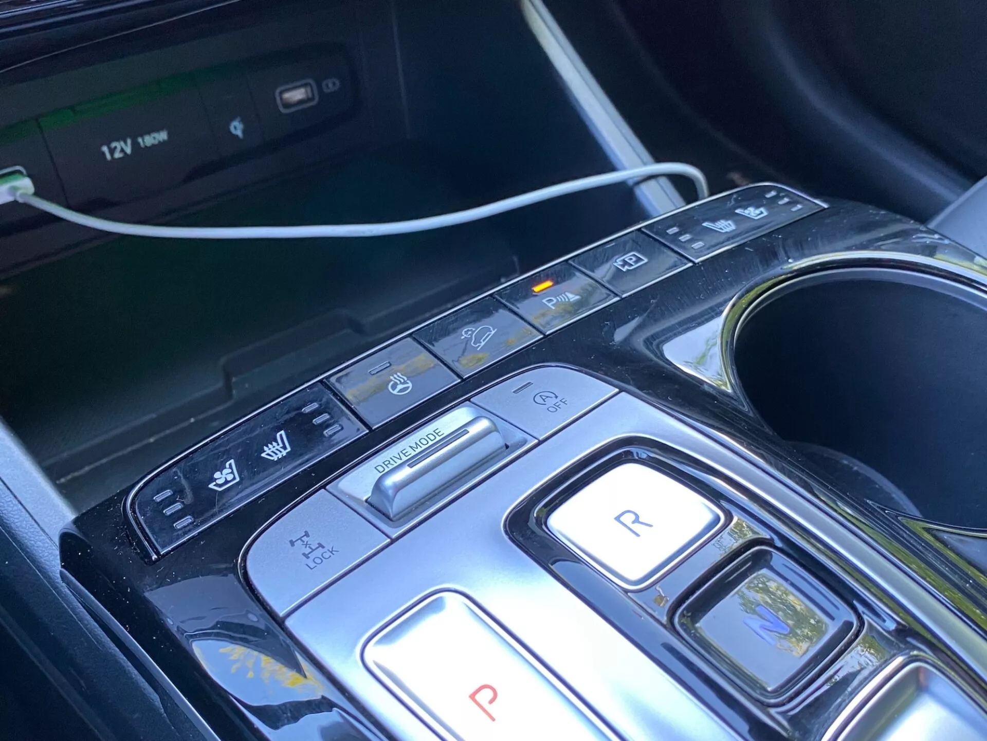 Detail středové konzole a výběru převodů vozu Hyundai Tucson 2020 1.6 T-GDi Pure, moderní design s tlačítky pro parkování, zpátečku, neutrál, jízdu a brzdění/regenerativní režimy, plus další tlačítka pro vypnutí automatického start/stop, parkovací senzory/automatické parkování a varování před opuštěním jízdního pruhu. Viditelný multimediální systém, držáky na nápoje a 12V zásuvka naznačují komfort, komfort a pokročilé technologie asistence řidiče (ADAS). Konzole má elegantní design s lesklým černým materiálem a kovovými akcenty, což naznačuje luxusní vzhled.