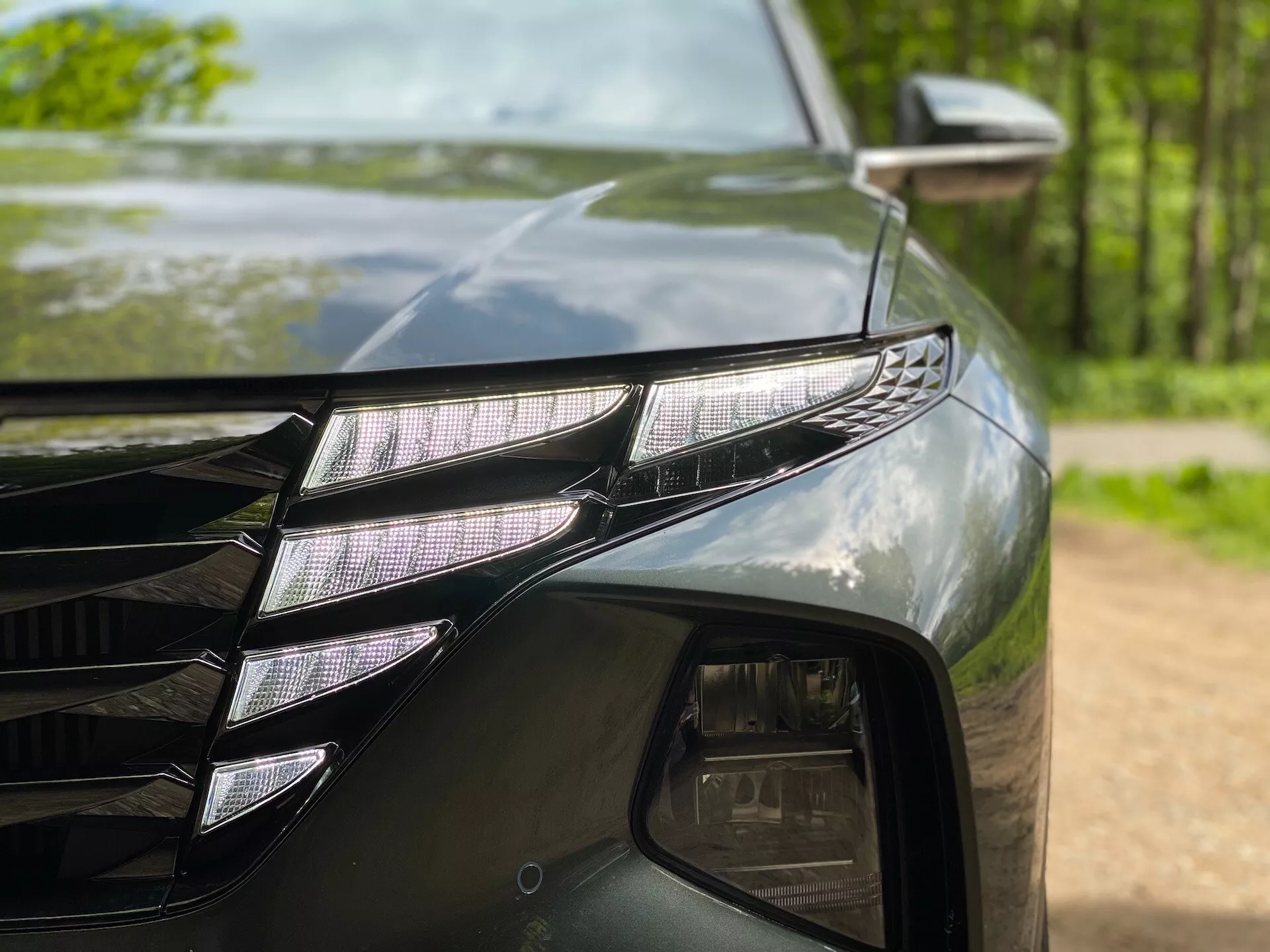 Detailní záběr přední levé části Hyundai Tucson 2020 (Hyundai-Tucson-1-6-T-GDi-Pure), ukazující LED světlomet s jednotlivými LED projektory a výrazným LED DRL proužkem. Tmavý, lesklý lak odráží okolní stromy. Viditelná je také část mřížky s hranatými liniemi a boční zrcátko s indikátorem směru.