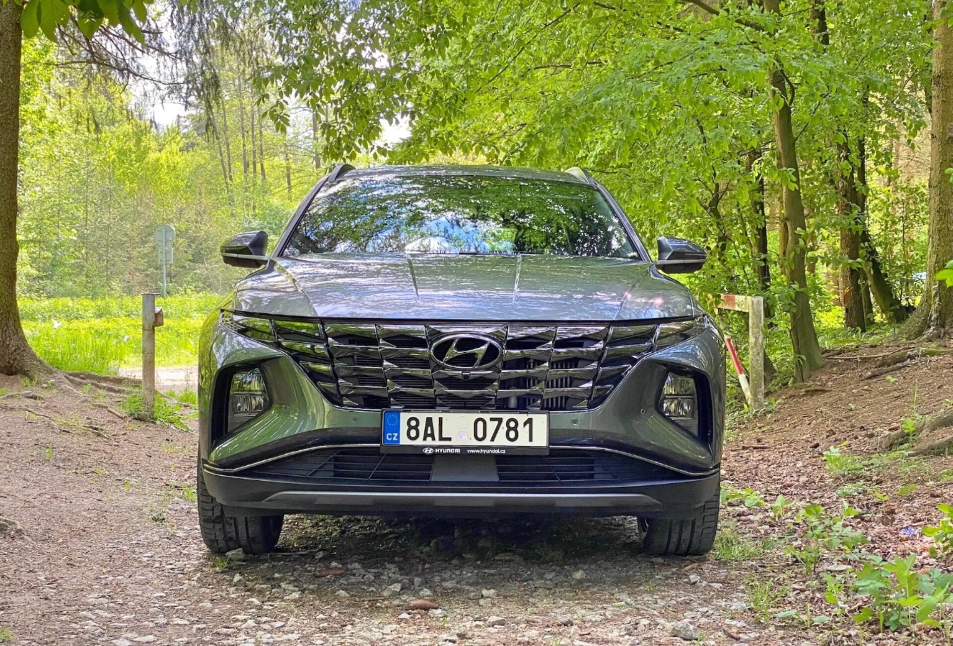 Přední pohled na vůz Hyundai Tucson 1.6 T-GDi Pure z roku 2020, zobrazující nízkoúhlý snímek zaměřený na mřížku, přední nárazník a oblast kapoty. Automobil je zaparkován na štěrkové cestě v přirozeném prostředí, což naznačuje jeho vhodnost pro venkovní prostředí. Klíčové vlastnosti zahrnují výrazný design mřížky, elegantní LED světlomety a viditelný znak Hyundai.