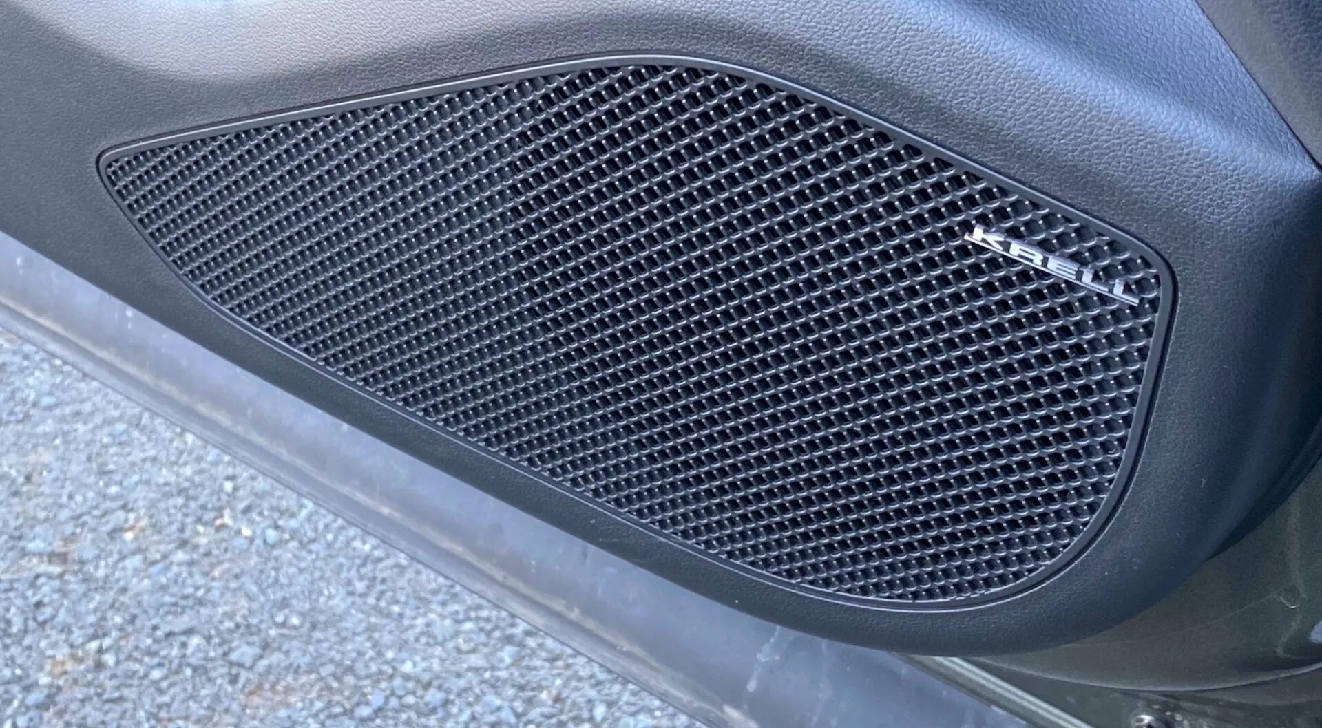 Detailní boční pohled na Hyundai Tucson 2020 1.6 T-GDI Pure, zaměřený na boční mřížku s diamantovým vzorem síťoviny. Mřížka, zapuštěná do tvarované kontury, je zakomponovaná do karoserie vozu s matným povrchem, obklopena hladkým zakřiveným povrchem. Obrázek, focený z úhlu zdola nahoru, ukazuje přirozené venkovní osvětlení s kousky asfaltu a silničními nečistotami, které podtrhují elegantní, výkonný vzhled.