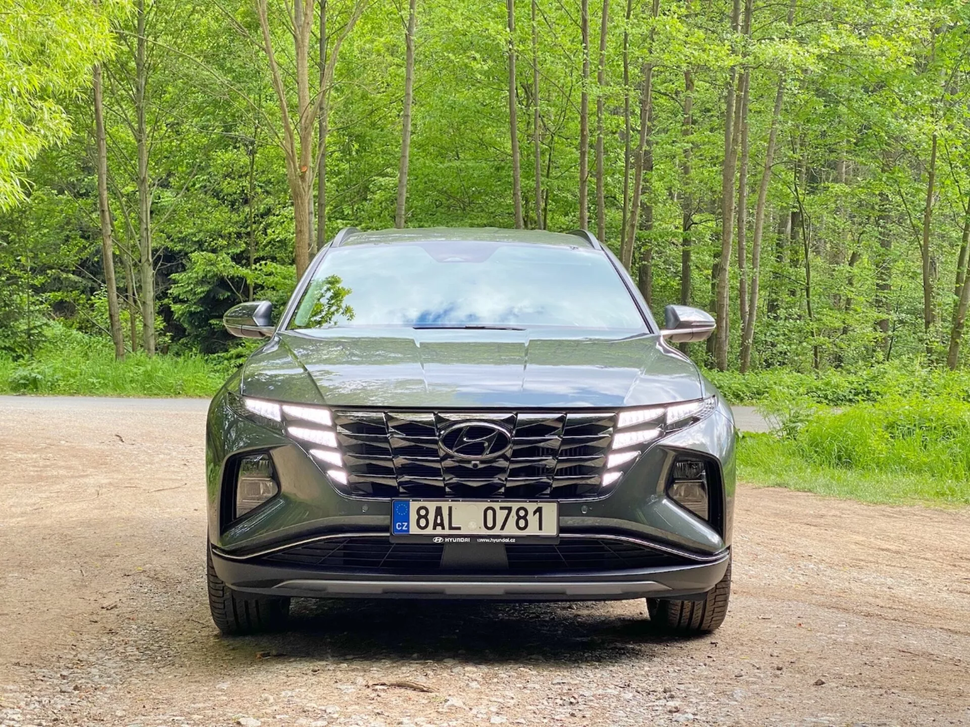 Čelní pohled na Hyundai Tucson 2020 (Hyundai Tucson 1.6 T-GDI Pure) moderní SUV zaparkované na štěrkové cestě s lesnatým pozadím. Obrázek zachycuje elegantní, agresivní design s velkou geometrickou mřížkou s logem výrobce, úzkými úhlovými LED světlomety a tvarovaným předním nárazníkem s nasávacími otvory a prostorem pro mlhové světlomety, zdůrazňující pokročilou aerodynamiku a osvětlovací technologii.