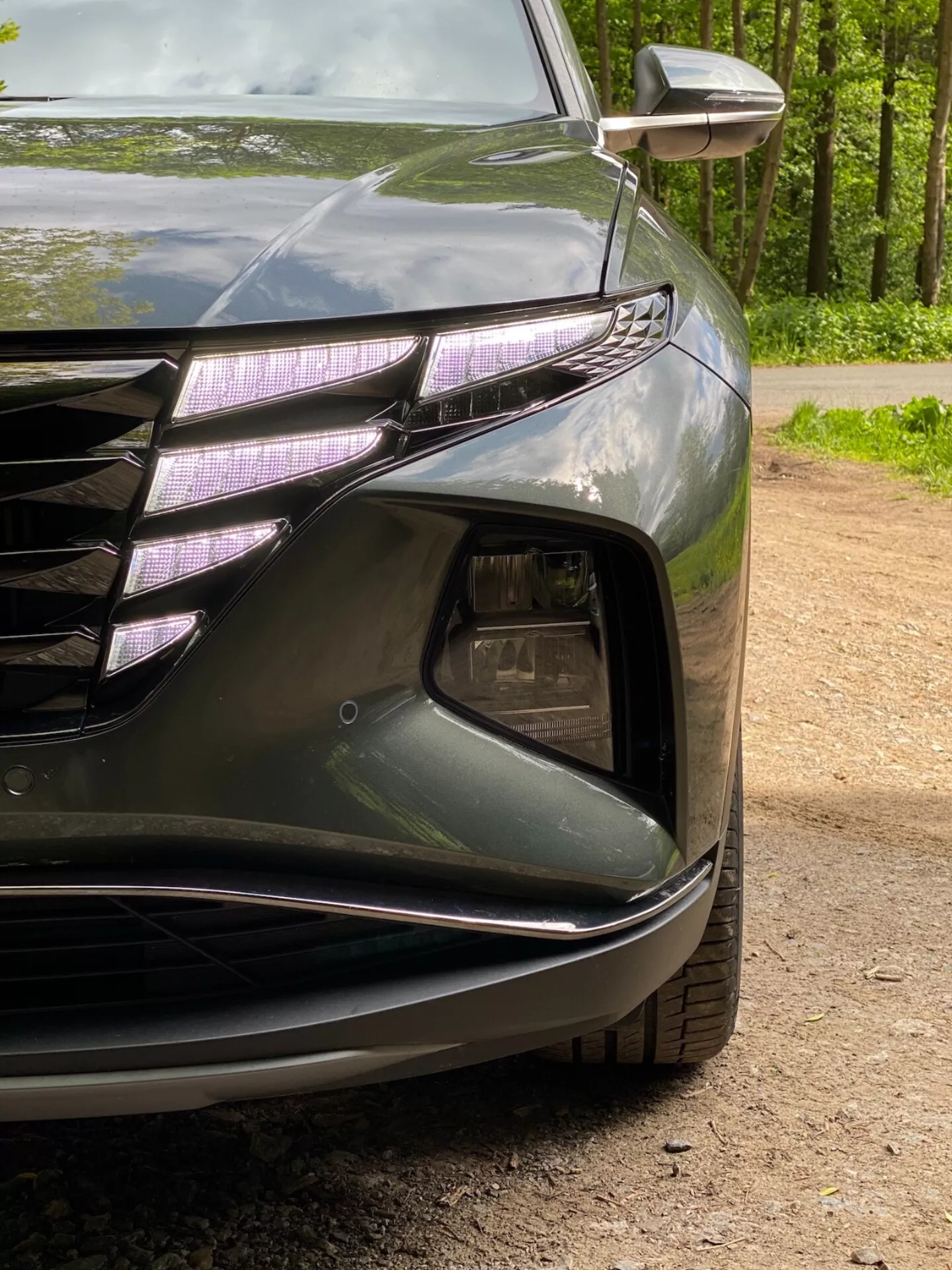 Detailní pohled na levý přední roh vozu Hyundai Tucson 2020 1.6 T-GDI Pure, zobrazující světlomet s LED denními světly a čočkou, část přední mřížky s horizontálními liniemi a chromovanými akcenty, sekci přívodu vzduchu s pouzdrem mlhovek, tvarovaný přední nárazník s hladkými liniemi a integrovaným směrovým světlem v zrcátku. Tmavý lesklý lak zdůrazňuje aerodynamické křivky vozu.