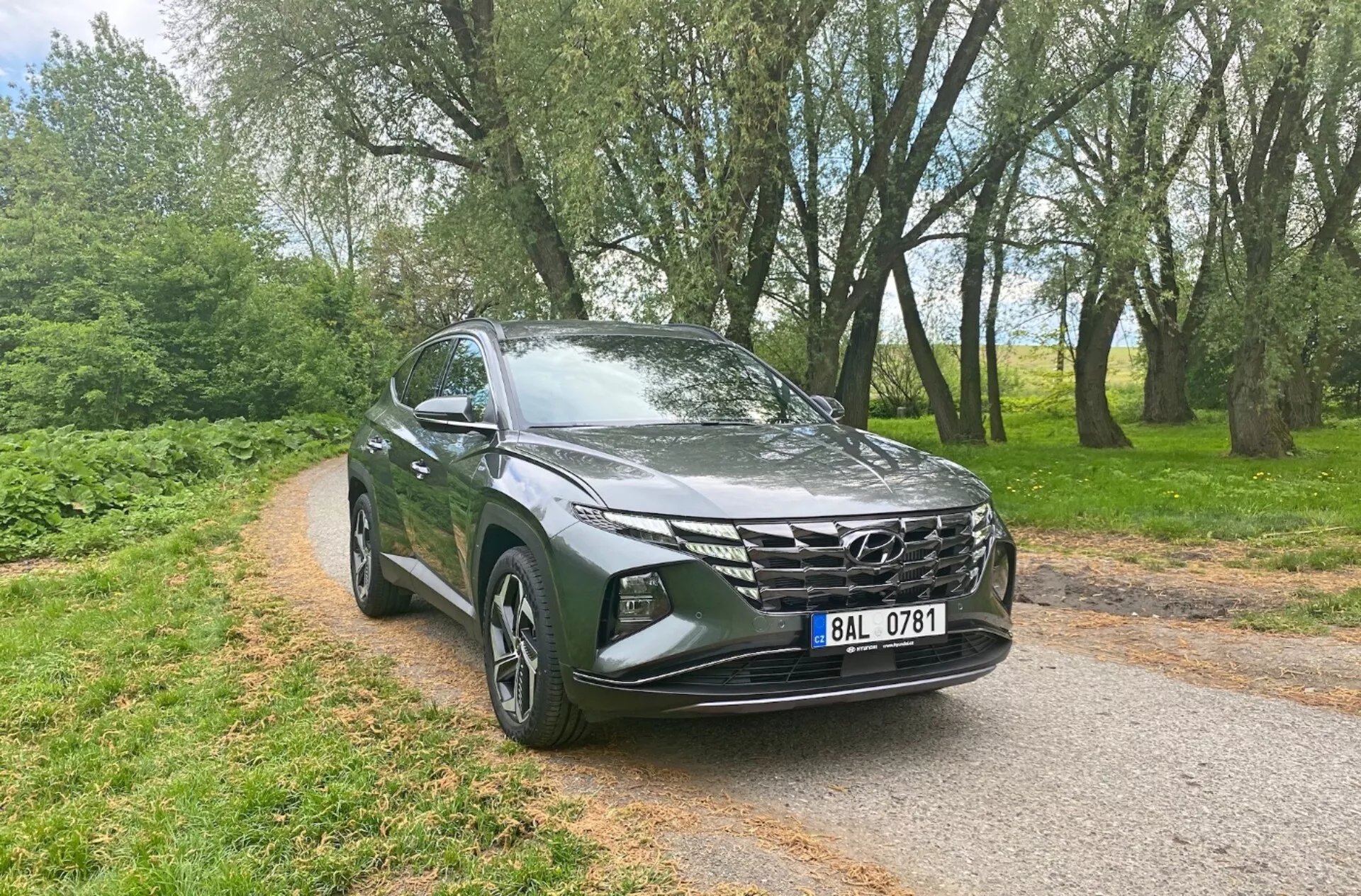 Tříčtvrteční pohled zepředu na Hyundai Tucson 1.6 T-GDi Pure 2020, ukazující moderní SUV prvky jako elegantní LED světlomety, výrazná geometrická maska, aerodynamická kapota, litá kola, zrcátka s integrovanými směrovými světly a metalízu, na pozadí stromů a úzké asfaltové silnice, což zdůrazňuje jeho všestrannost pro městské i venkovské prostředí.