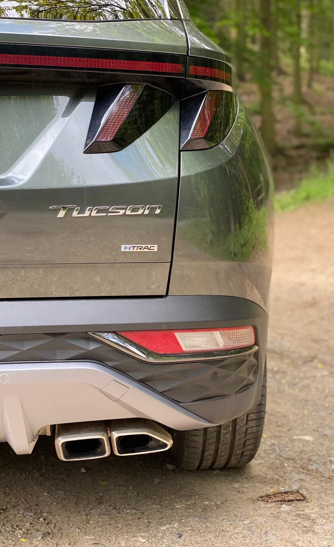 Částečný zadní pohled na Hyundai Tucson 2020 model, který naznačuje, že se jedná o verzi 1.6 T-GDi Pure, zobrazující pravou stranu nárazníku, částečné zadní světlo, zadní spojler s LED brzdovým světlem, dvojité výfukové koncovky a označení modelu s názvem 'Tucson' a technologií motoru 'T-GDi'.