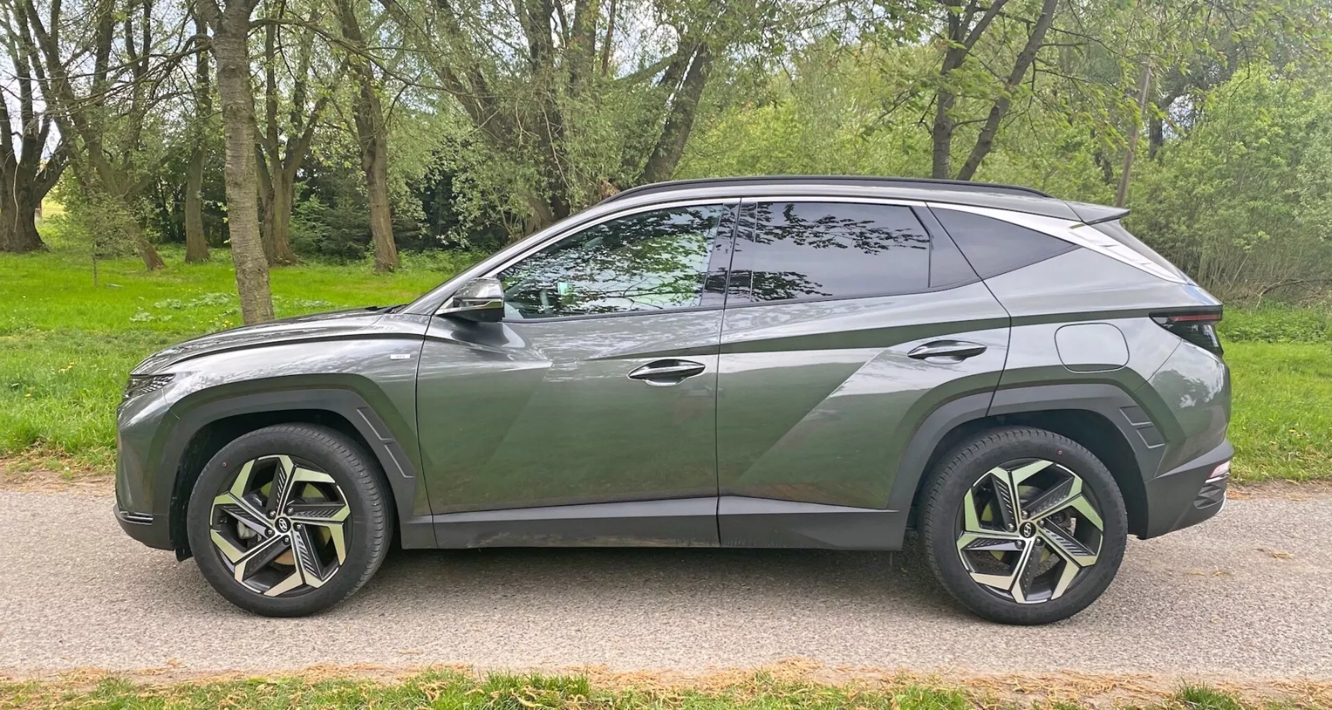Boční profil moderního SUV Hyundai Tucson 1.6 T-GDI Pure z roku 2020 zaparkovaného na dlážděné silnici vedle travnaté plochy s stromy. Představuje elegantní, aerodynamický design se svažující se střechou, výraznou linií karoserie, velkými vícepaprskovými litými koly s pneumatikami s nízkým profilem, tmavými okny s viditelným B-sloupkem, zapuštěnými klikami dveří, barevnými bočními zrcátky a chromovaným lemováním podél okna. Vozidlo zachycuje moderní styl a možné pokročilé funkce z bočního úhlu.