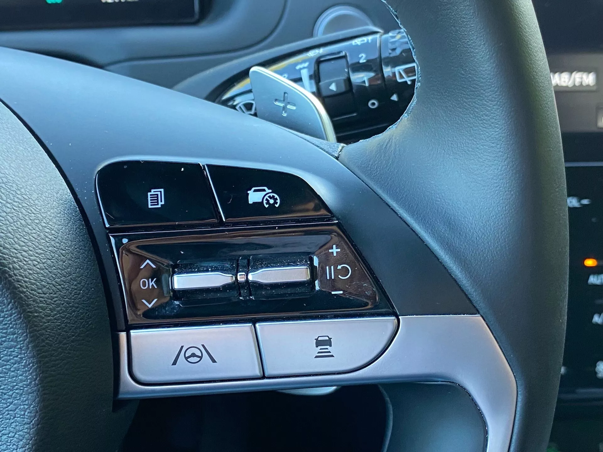 Detailní pohled na ovládací prvky na volantu na levé straně Hyundai Tucson 1.6 T-GDI Pure z roku 2020, ukazující funkce jako navigace v menu, ovládání hlasitosti audia, aktivace hlasového příkazu a funkce Bluetooth pro telefonování.