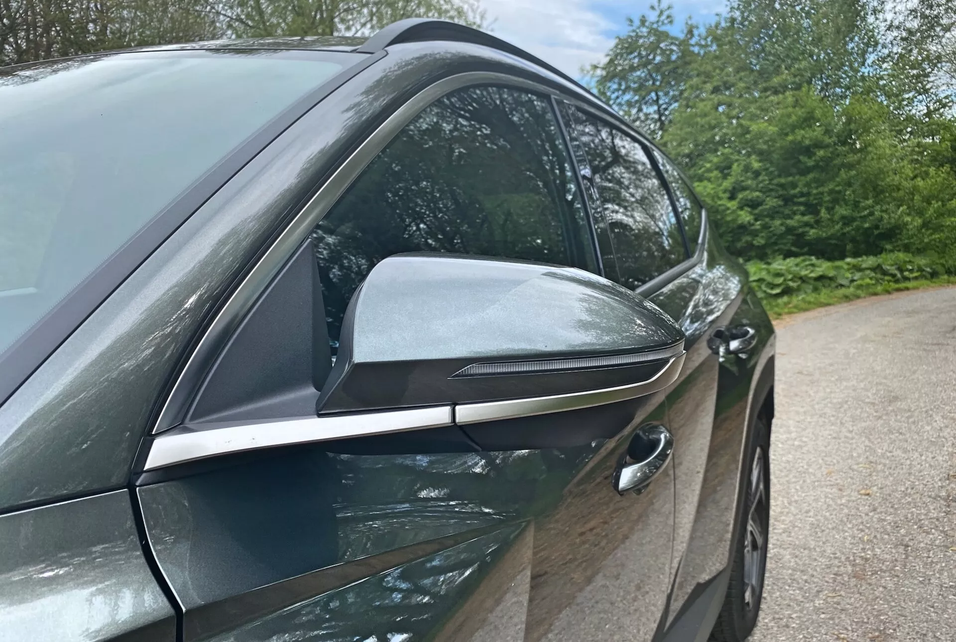 Detailní pohled z boku na moderní design a technologické prvky Hyundai Tucson 2020 1.6 T-GDI Pure se zaměřením na stranu řidiče. Fotografie z nízkého úhlu zdůrazňuje elegantní, aerodynamicky tvarované boční zrcátko s integrovanými směrovkami a černými akcenty, což naznačuje pokročilé funkce. Zapustěná přední klika dveří řidiče naznačuje bezklíčový vstup, čímž přispívá k aerodynamické účinnosti vozidla. Lesklé, tělově zbarvené dveře a okenní lišty s chromem naznačují prémiový balíček výbavy. Pozadí s přírodními prvky jako stromy a obloha naznačuje venkovní prostředí.