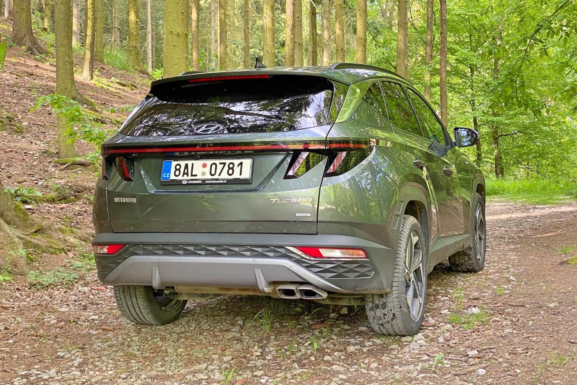 Obrázek moderního SUV Hyundai Tucson 2020 v lesním prostředí na štěrkové cestě, zachycený z pravého zadního tříčtvrtečního úhlu. Fotografie zdůrazňuje pravou stranu a zadní profil vozidla s viditelnými zadními dveřmi, LED zadními světly, dvojitým výfukovým systémem, zadním spoilerem a hliníkovými koly.