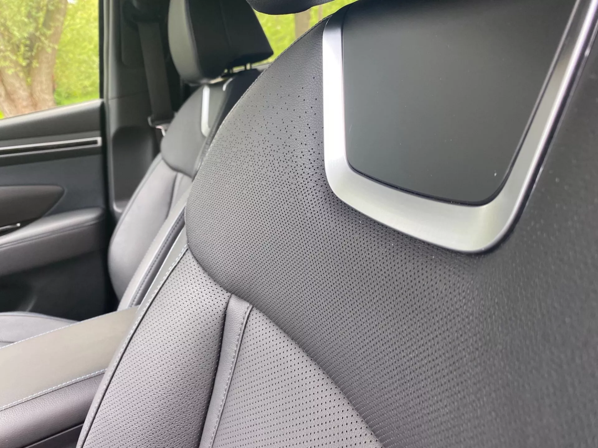 Přiblížený záběr interiéru vozu Hyundai Tucson 2020, zaměřený na pravý A-sloupek a část pravého předního sedadla. A-sloupek je pokryt texturovaným materiálem, pravděpodobně látkou nebo syntetickou kůží, což naznačuje estetické nebo redukční zaměření na hluk. Sedadlo má dvoubarevný design a je perforované pro větrání. Výrazný prvek na křižovatce A-sloupku a stropní výplně by mohl být součástí audio systému, komponentem airbagu nebo senzorem asistence řidiče, zdůrazňující pokročilé bezpečnostní nebo technologické funkce.