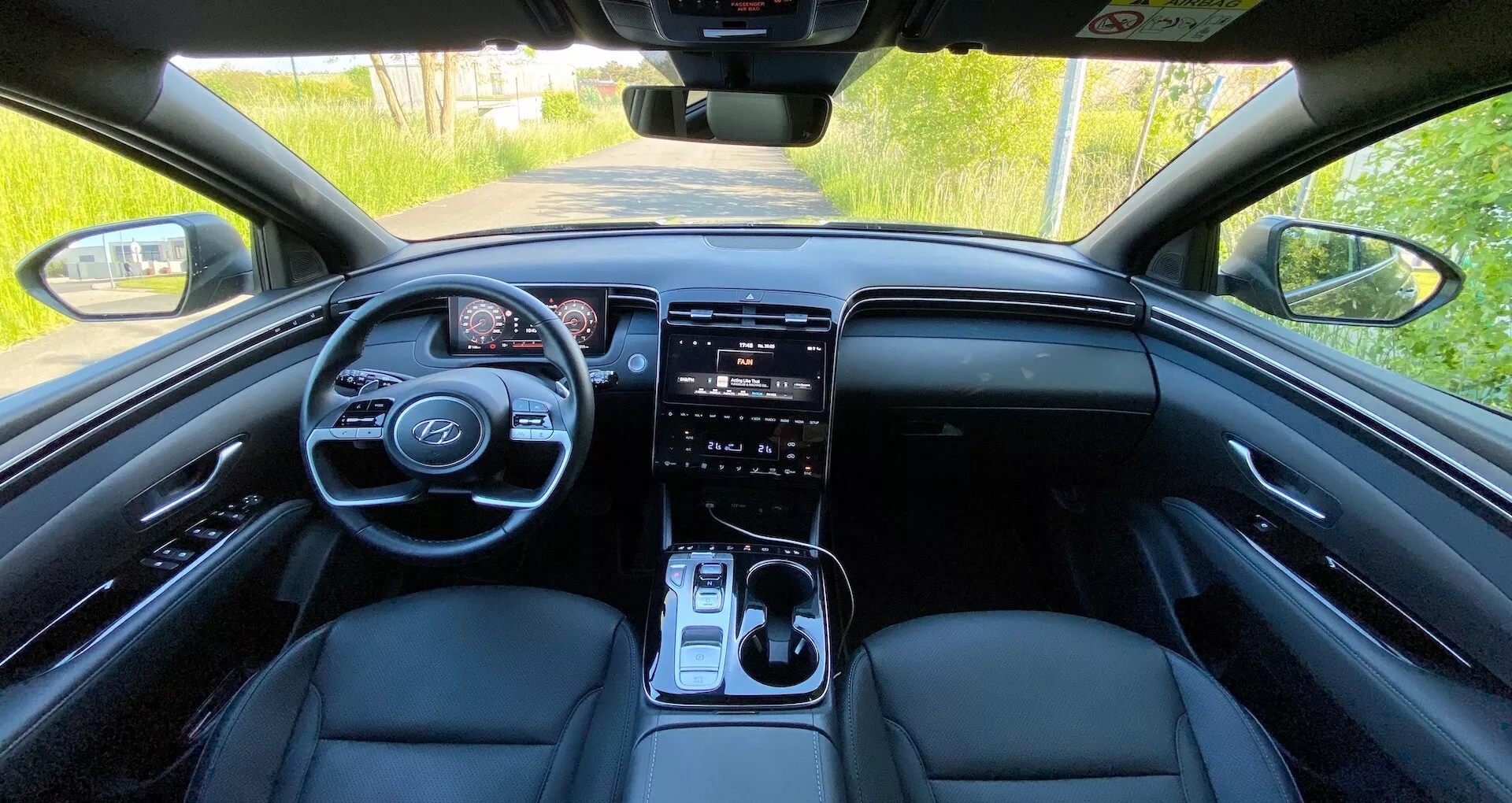 Interiér Hyundai Tucson 2020 z pohledu zadního sedadla s pohledem na přední palubní desku, volant s ovládacími prvky, digitální přístrojový panel, velký dotykový informační systém, tlačítka pro ovládání klimatizace, řadicí páku a přední sedadla potažená kůží za přirozeného denního osvětlení.