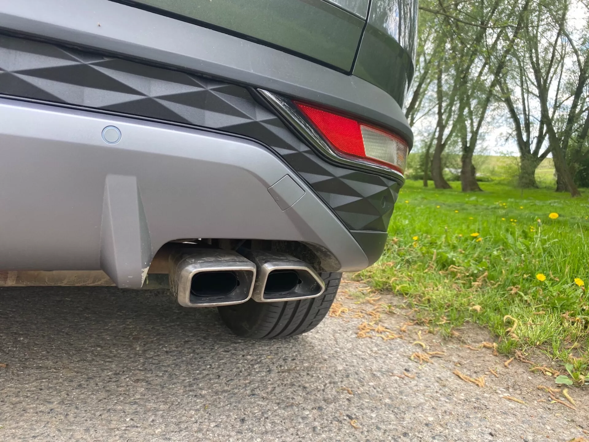 Detailní záběr zadního rohu vozu Hyundai Tucson 2020 z úrovně země. Viditelné detaily zahrnují dvoubarevný zadní nárazník, částečné LED zadní světlo a dvojité kovové výfukové trubky s obdélníkovými zakončeními, což naznačuje sportovní výbavu. Design vozu je elegantní a metalický lak odráží okolní prostředí.