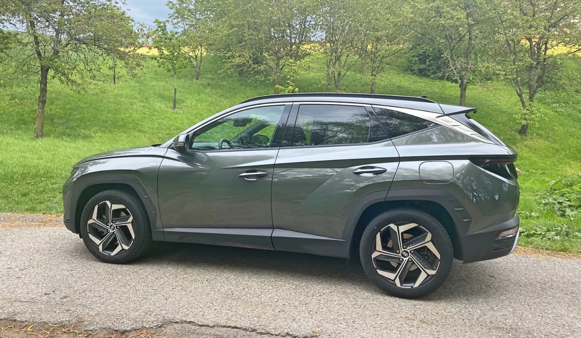Boční profil 2020 Hyundai Tucson 1.6 T-GDI Pure SUV v přírodním denním světle. Vozidlo se nachází na travnaté ploše s stromy v pozadí, což naznačuje venkovské nebo předměstské prostředí. Moderní designové prvky zahrnují LED světlomety, integrované směrovky na bočních zrcátkách, tónovaná okna, aerodynamická hliníková kola a klesající střechu jako u kupé. Elegantní karoserie s charakterovými liniemi a zapuštěnými klikami dveří naznačují pokročilý systém bezklíčového vstupu, zatímco zadní spojler naznačuje aerodynamická vylepšení. Auto je centrálně umístěno v rámečku a poskytuje čistý, nebráněný pohled ze střední výšky.