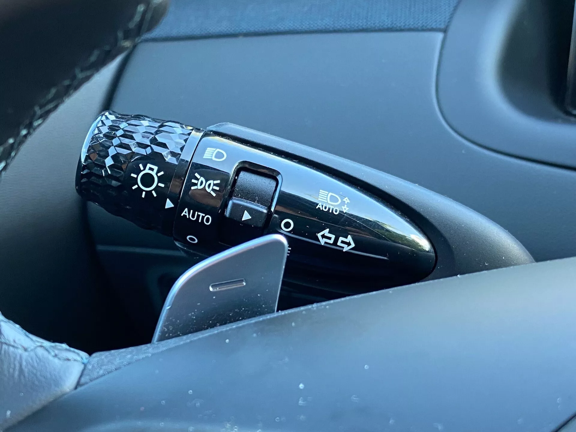 Detailní pohled na ovládací páku umístěnou na sloupku řízení nalevo od volantu v Hyundai Tucson 2020. Páka obsahuje symboly pro nastavení světlometů včetně vypnutí, automatických světlometů, parkovacích světel a potkávacích/dálkových světel. Obrázek zachycuje páku, část volantu a rozmazané pozadí řidičových dveří, zdůrazňující detail a kvalitu designu. Poloha 'AUTO' indikuje automatický systém světlometů.