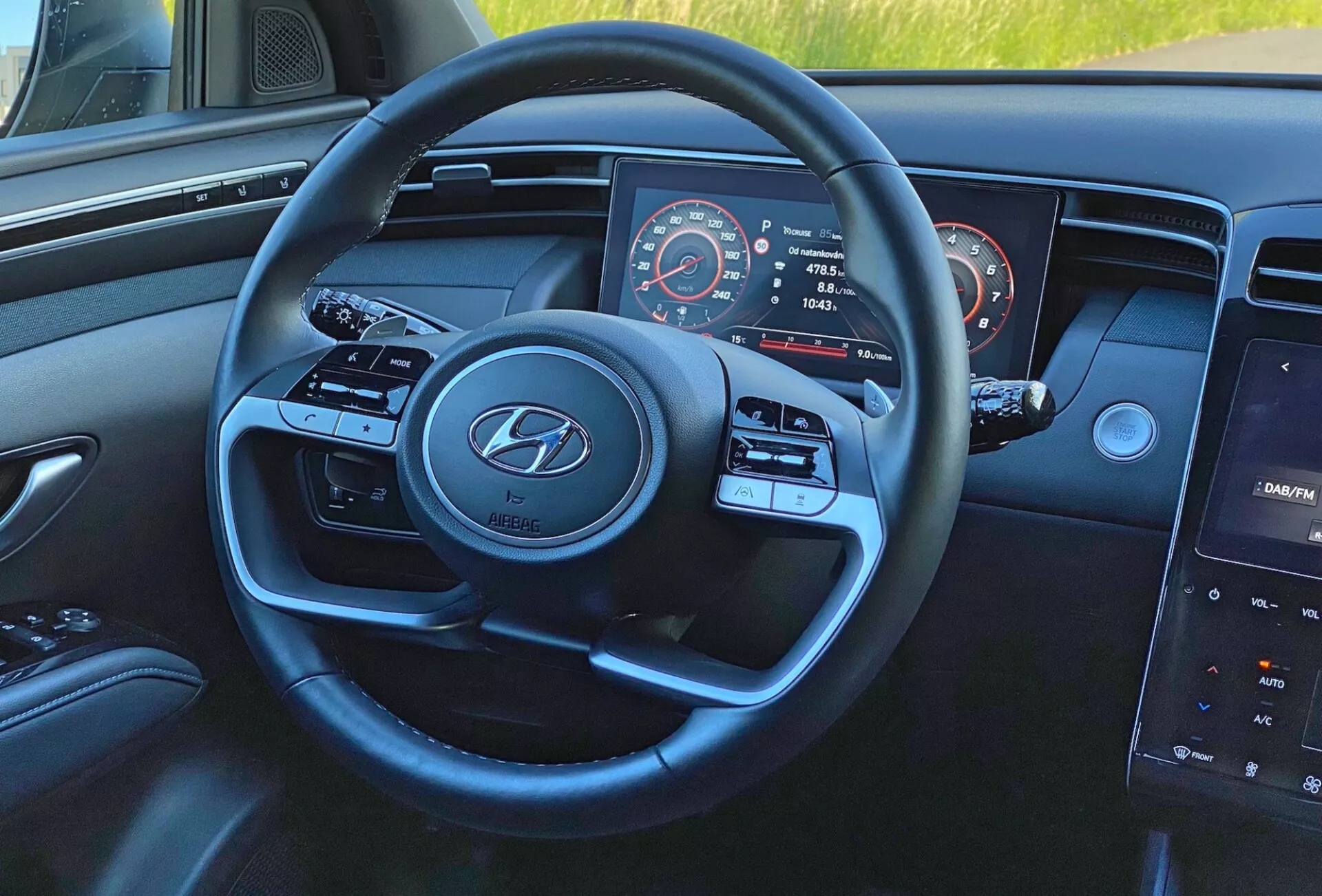 Interiér vozu Hyundai Tucson 1.6 T-GDI 2020 z pohledu řidiče se zaměřením na moderní tříramenný volant s logem Hyundai, tlačítka pro média a tempomat, částečně viditelný přístrojový panel s tachometrem a otáčkoměrem a centrální dotykový infotainment systém na palubní desce.