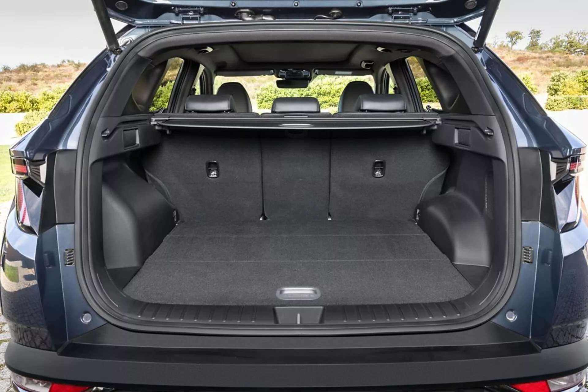 Zadní nákladový prostor Hyundai Tucson 2020 s otevřeným kufr, předvádějící kobercem pokrytou nákladní plochu, body pro upevnění, úložné přihrádky, 12V zásuvku, skládací zadní sedadla, zasunutý kryt nákladu, odolné plastové bočnice a pohled na zadní nárazník, v exteriéru.