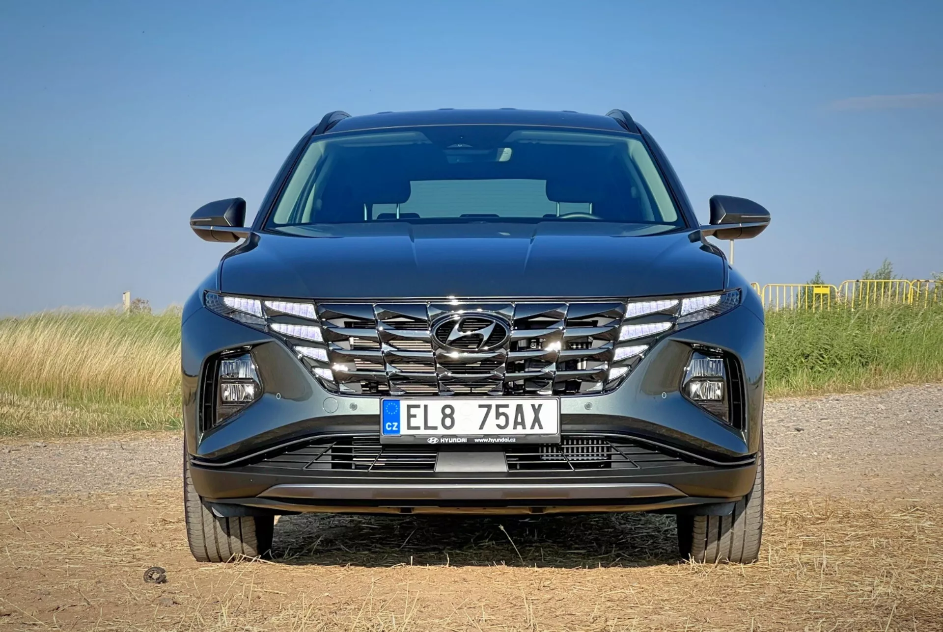 Pohled zepředu na Hyundai Tucson 2020 předvádějící aerodynamický design kapoty, kaskádovou masku chladiče, ostré LED světlomety a detailní nárazník s možnými technologickými prvky, umístěný na pozadí přírodní krajiny