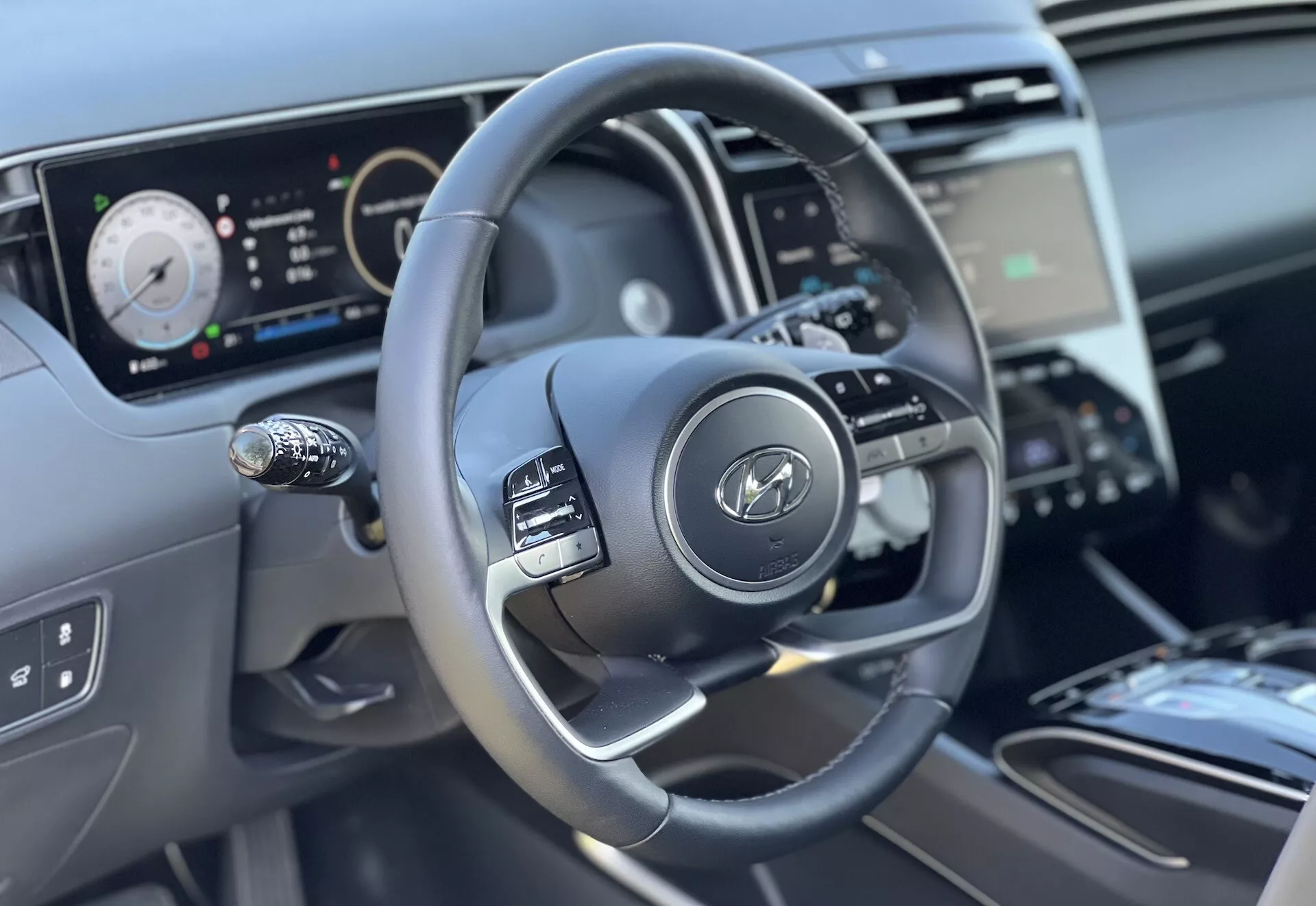 Vnitřní pohled na Hyundai Tucson 2020 se zaměřením na volant a prvky palubní desky z perspektivy řidiče, zdůrazněním tříramenného designu s ovládacími tlačítky, digitálního přístrojového panelu a částečného pohledu na středovou konzolu a ovládání klimatizace.