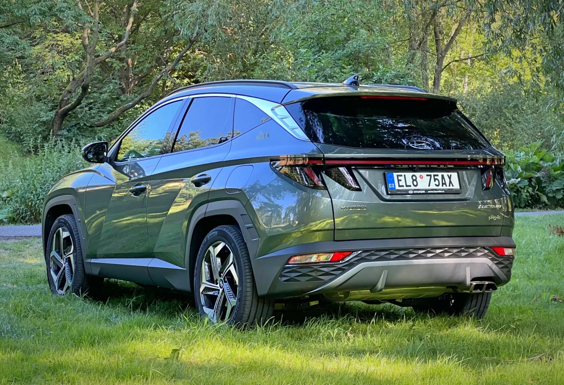 Zadní tříčtvrteční pohled na Hyundai Tucson 2020 zdůrazňující LED zadní světla, parkovací senzory, dvojitý výfuk, sportovní zadní spoiler a střešní nosiče, zaparkovaný venku na trávě, prezentující aerodynamický design a pokročilé funkce moderního crossover SUV pro dobrodružství v přírodě a praktické využití.
