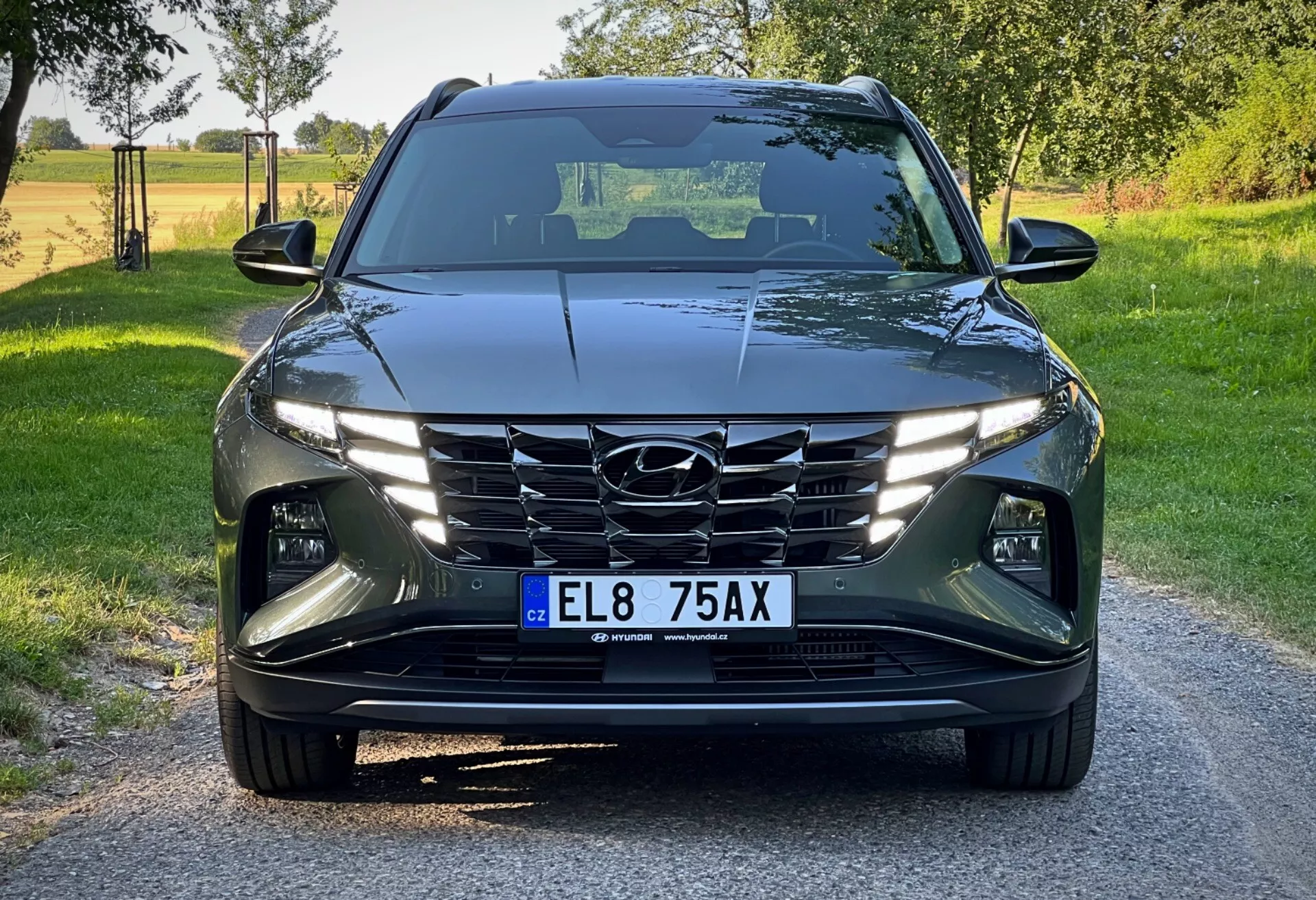 Hyundai Tucson 2020 zobrazen z přední perspektivy pod nízkým úhlem, prezentující jeho moderní design se sharp, úhlovými liniemi, LED světlomety, kaskádovou mřížkou a pokročilými systémy asistence řidiče na pozadí jasného venkovského dne