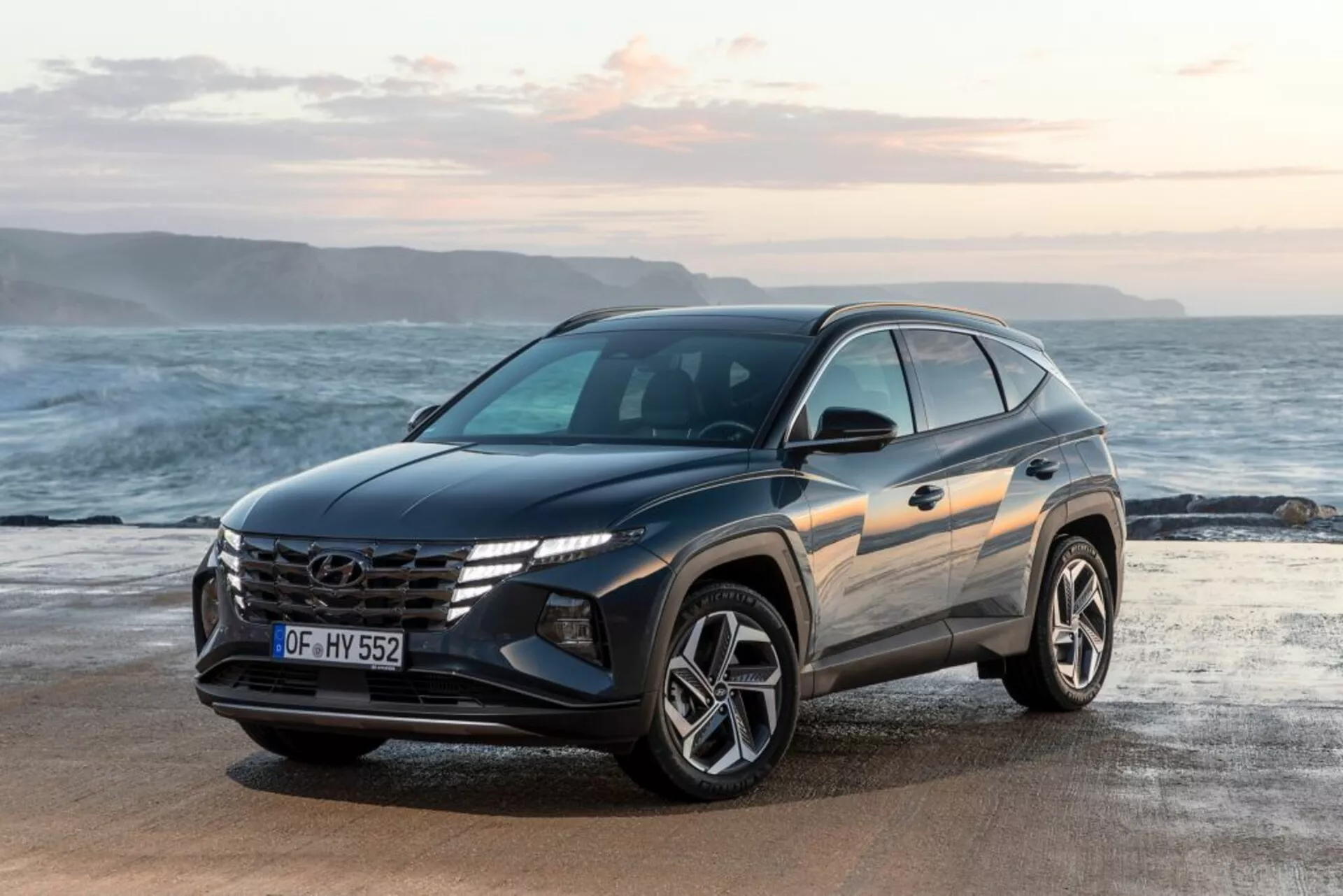 Moderní SUV Hyundai Tucson 2020 zaparkované na pobřeží s pozadím oceánu a horizontu, zdůrazňující pohled z předního tříčtvrtečního úhlu, který upozorňuje na design vozidla, výraznou mřížku s logem, úhlová LED světla a sportovní slitinová kola
