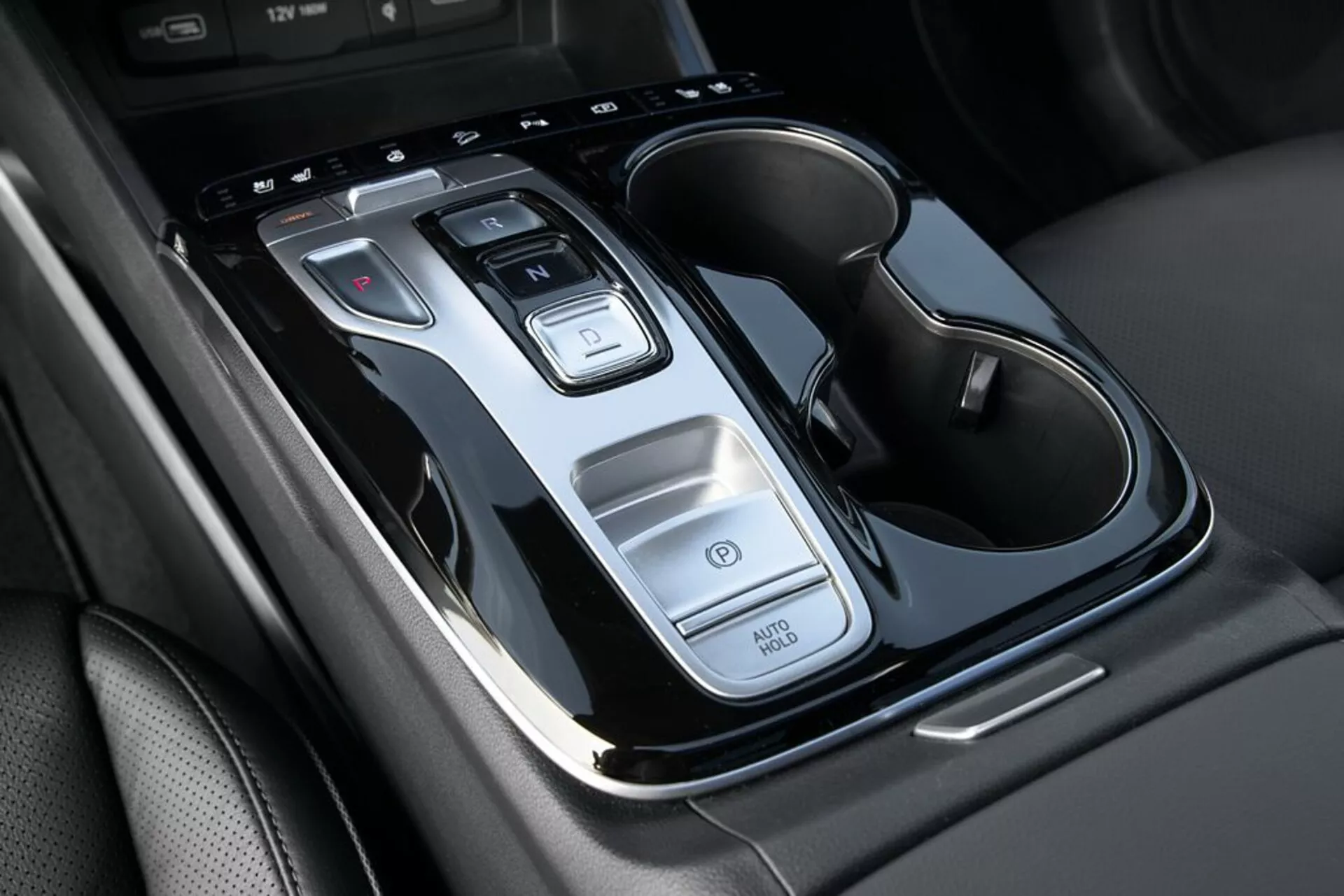 Detailní pohled na středovou konzoli Hyundai Tucson 2020 s pákou automatické převodovky, tlačítky ovládání, funkcí Auto Start/Stop, vypínačem výstražných světel a dvojitým držákem na nápoje s chromovými prvky, z pohledu shora s mírným sklonem.