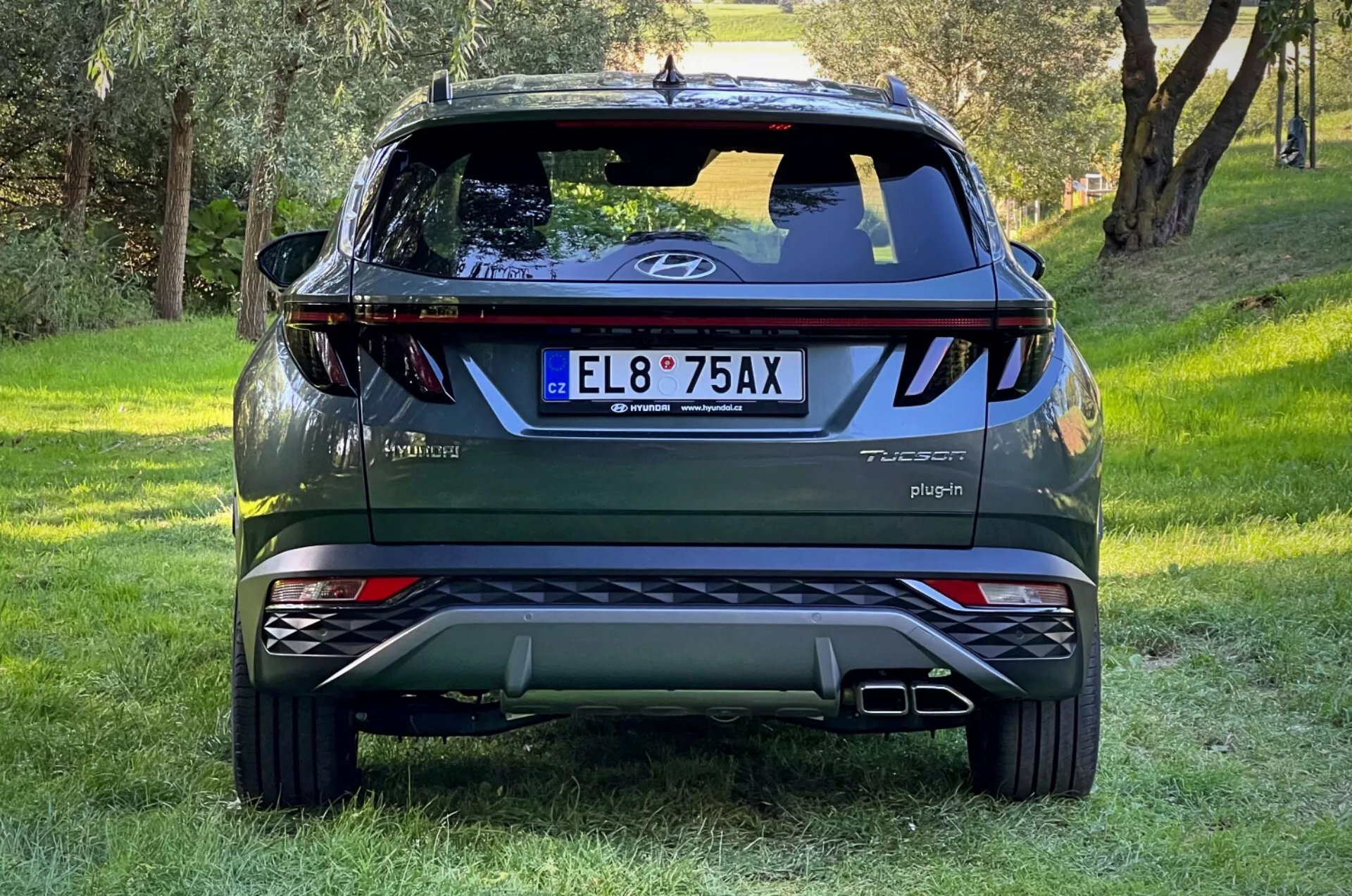 Zadní pohled na SUV Hyundai Tucson 2020 na travnatém svahu s lesním pozadím, ukazující logo na zavazadlovém prostoru, odznaky Tucson a Hybrid, nápis Plug-in, reflexní světla, dynamická zadní světla, zadní stěrač, vysoko umístěné brzdové světlo, dvojité výfukové špunty, stříbrné ochranné desky a odznak AWD.