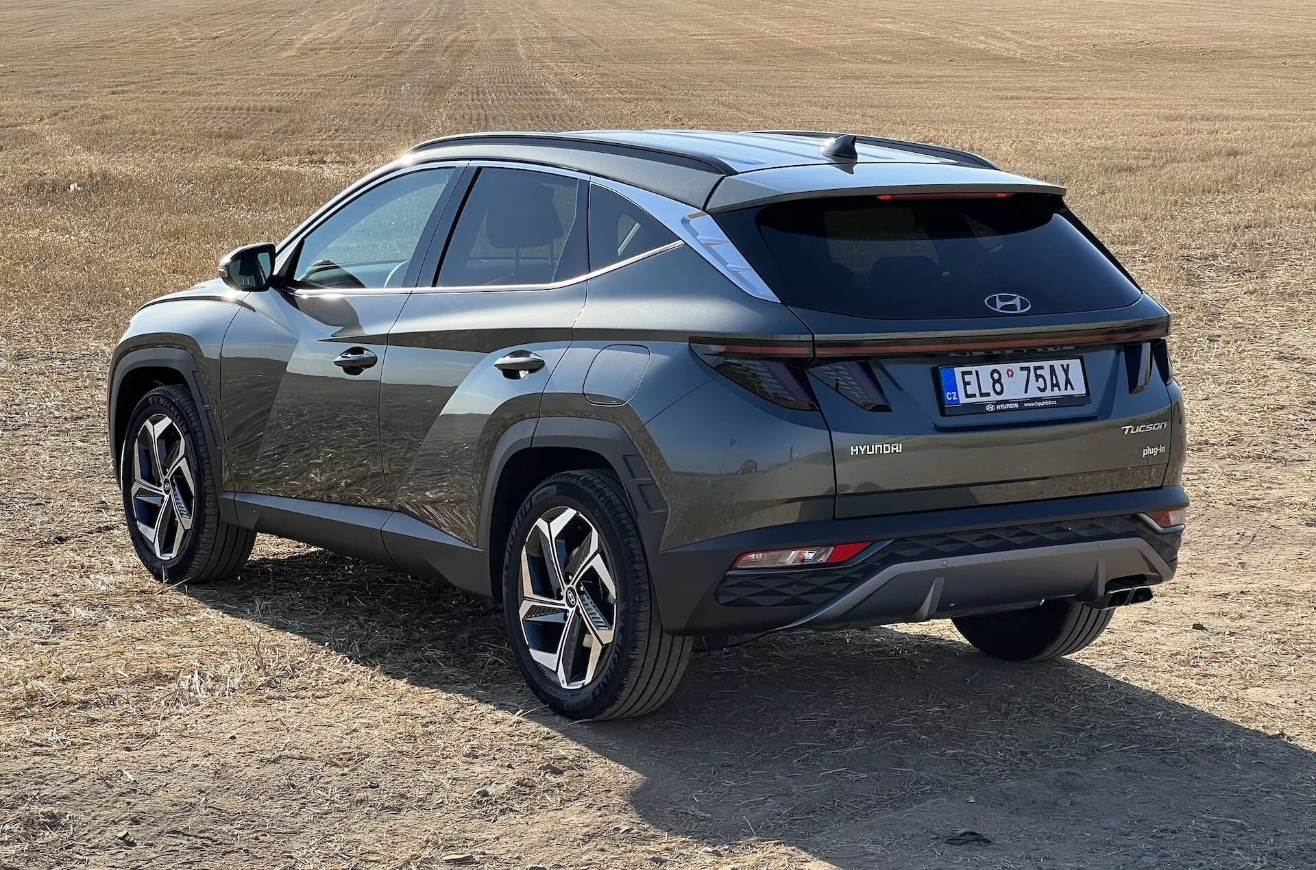 Moderní SUV Hyundai Tucson 2020 v šikmém pohledu ukazující zadní a boční profily s označením hybridní technologie na suchém travnatém poli, zdůrazňující LED zadní světla, kontrastní vložku nárazníku a sportovní kola s více paprsky