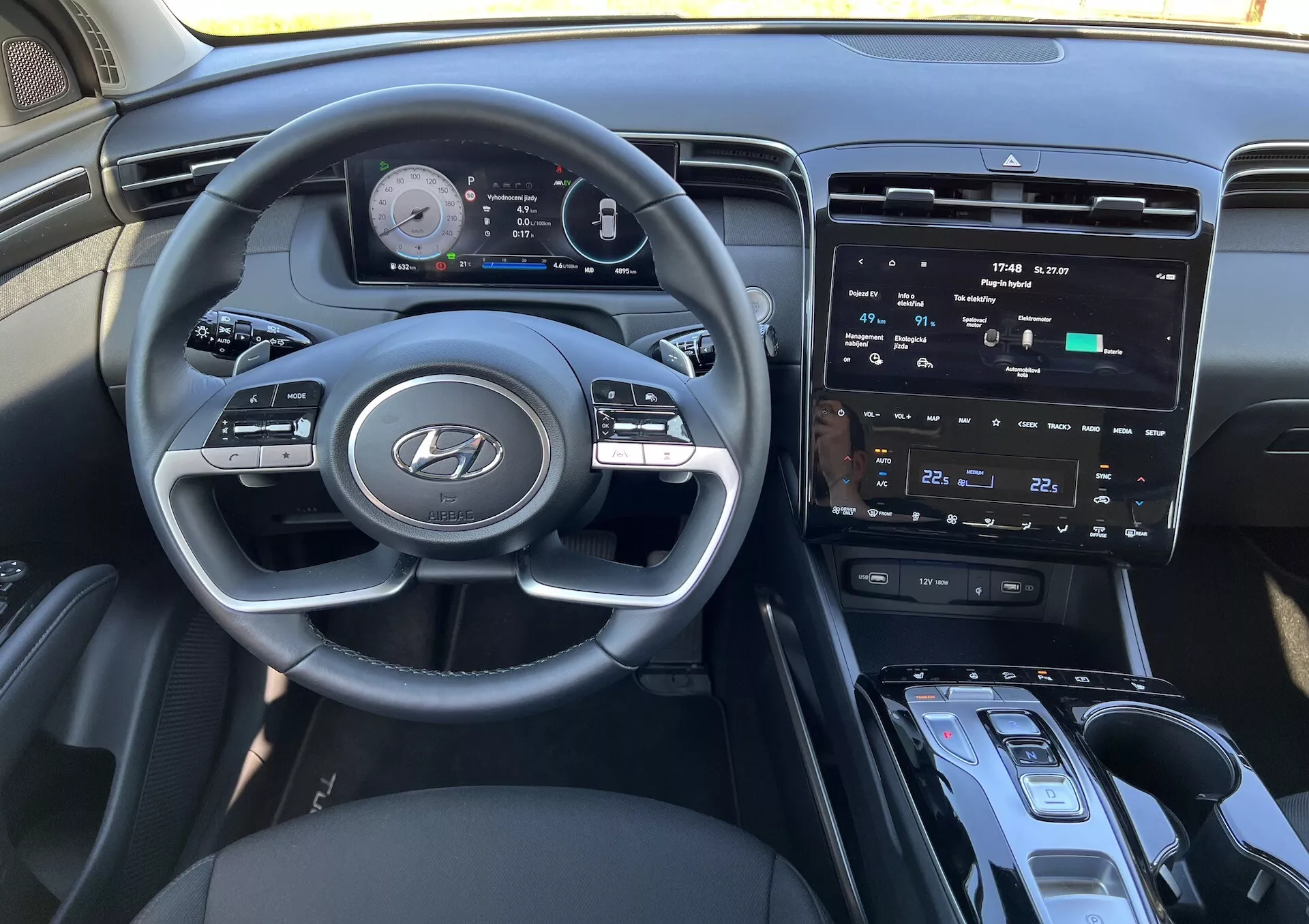 Interiér Hyundai Tucson 2020 s volantem s logem Hyundai, pokročilým digitálním přístrojovým panelem, centrálním dotykovým informačním systémem, ovládáním klimatizace, tlačítkem start/stop pro zapalování, pákou řazení a možnou oblastí pro bezdrátové nabíjení z pohledu řidiče