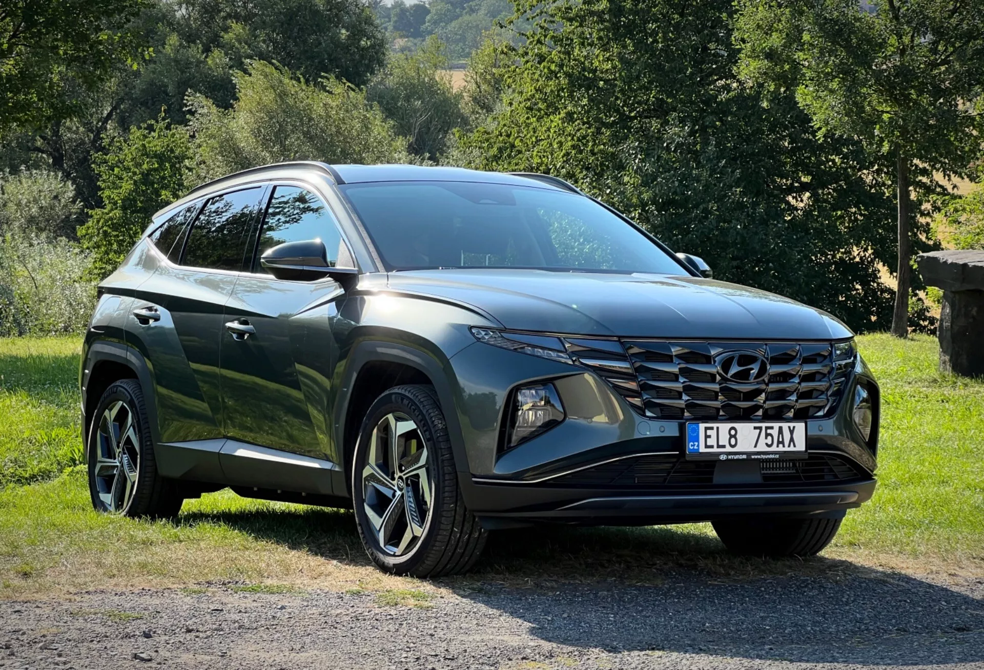 Hyundai Tucson 2020 zaparkovaný na trávě s venkovským pozadím, zobrazující perspektivu tří čtvrtin zepředu pro detailní pohled na jeho elegantní LED světlomety, úhlovou přední mřížku a lesklý tmavý lak