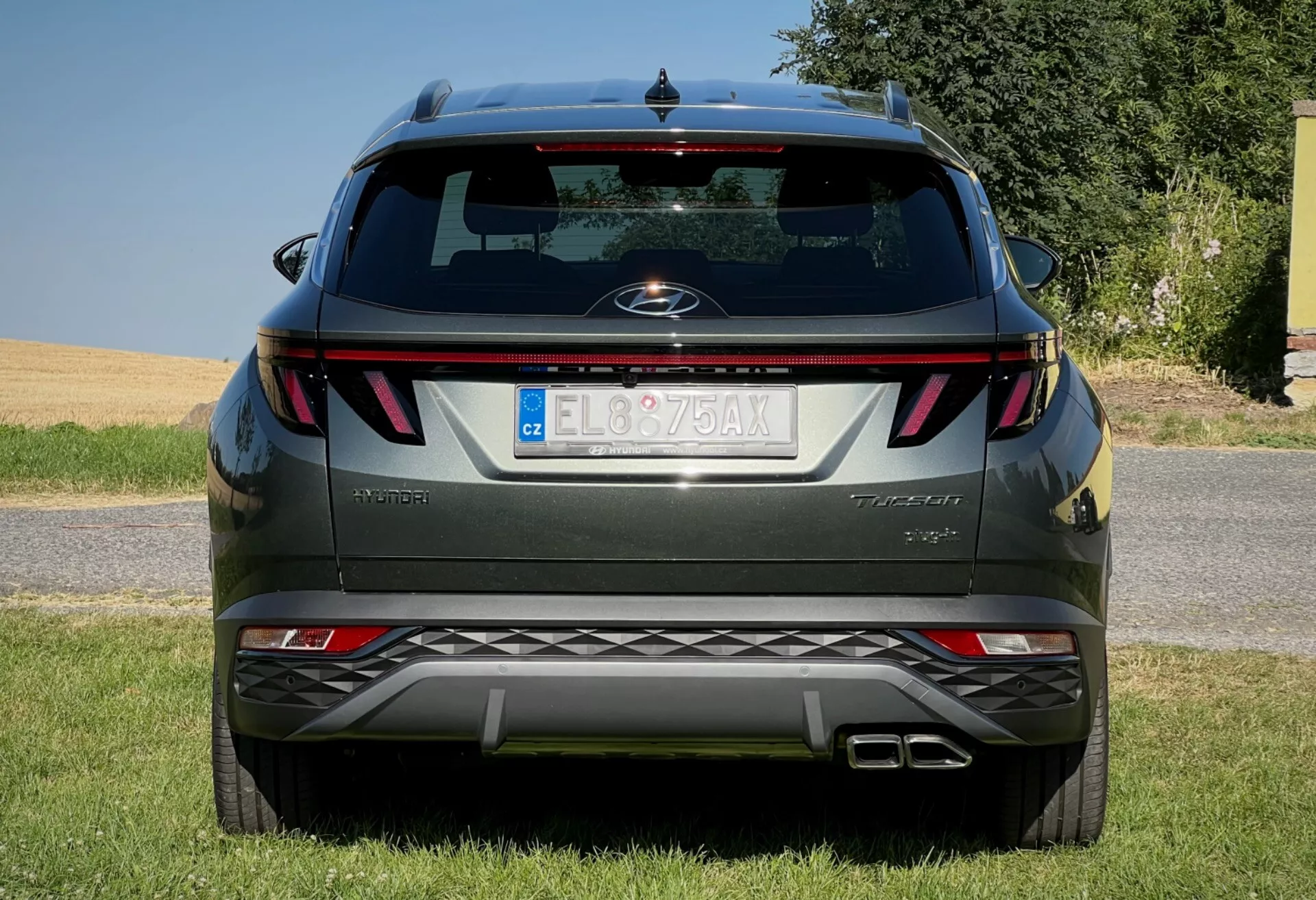 Zadní pohled na Hyundai Tucson 2020 ukazující horizontální zadní světla, centrální znak, název úrovně výbavy, integrované senzory parkování na nárazníku a dvojitý výfukový systém, zaparkovaný na ploše lemované trávou proti jasné obloze