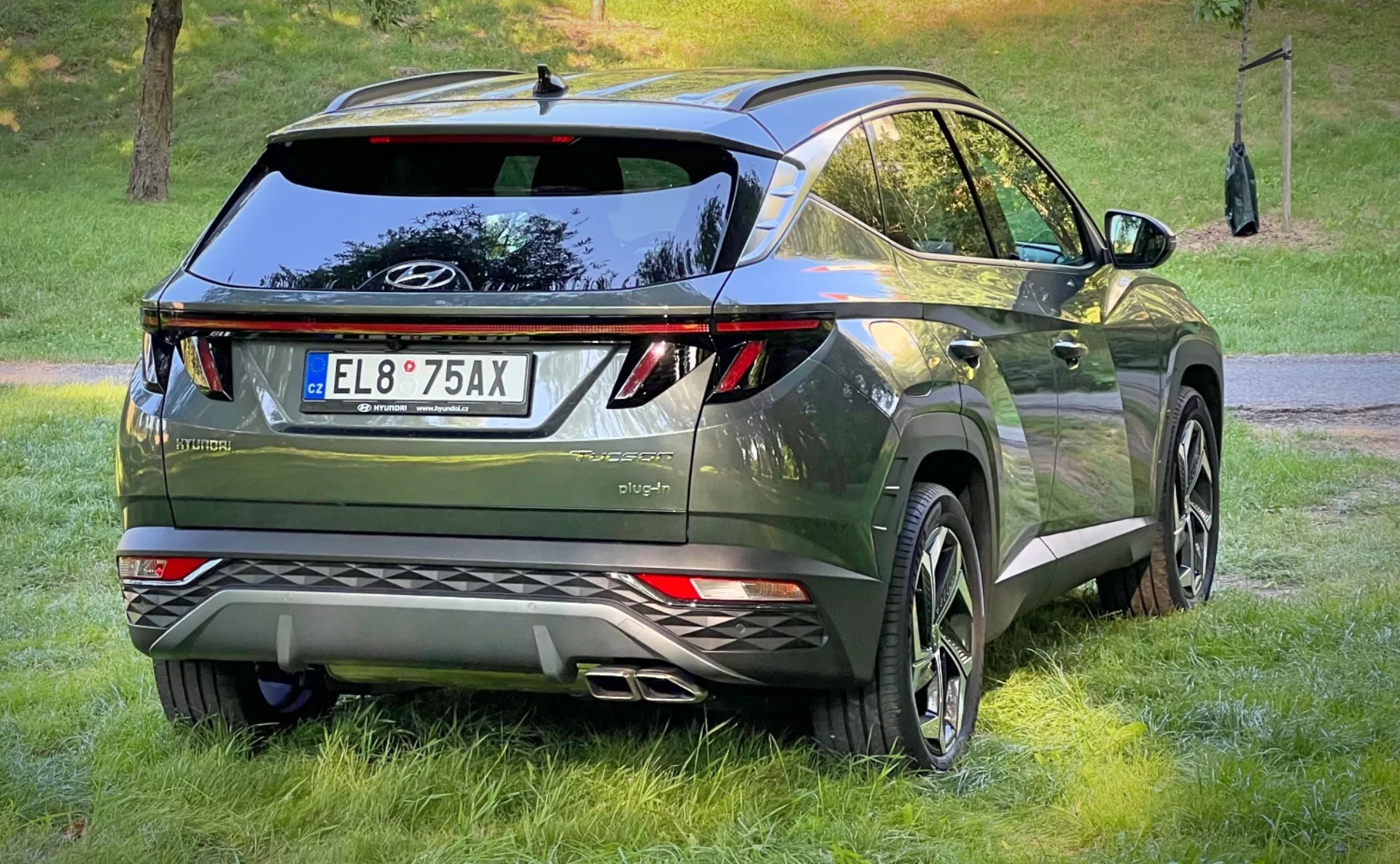 Zadní tříčtvrteční pohled na Hyundai Tucson 2020, předvádějící jeho LED zadní světla, aerodynamický design s křídlem na střeše a anténou ve tvaru žraločí ploutve, prémiový tmavě metalický zelený povrch a vybaven velkými slitinovými koly a parkovacími senzory v prostředí městského crossoveru