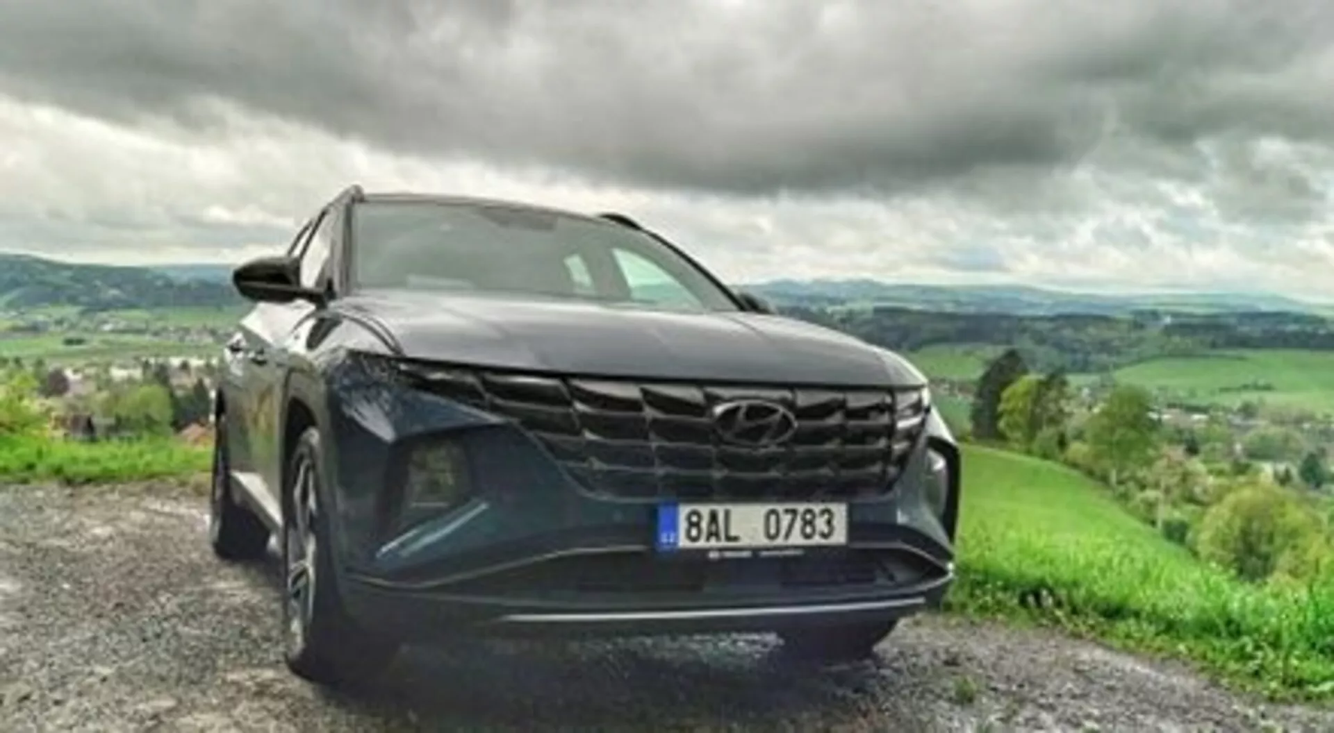 SUV Hyundai Tucson 2020 zaparkované na štěrkové cestě, předvádí svou odvážnou mřížku, štíhlé LED světlomety a metalický lak v dynamickém pohledu z nízkého předního úhlu, s bujnou kopcovitou krajinou a zataženým nebem na pozadí